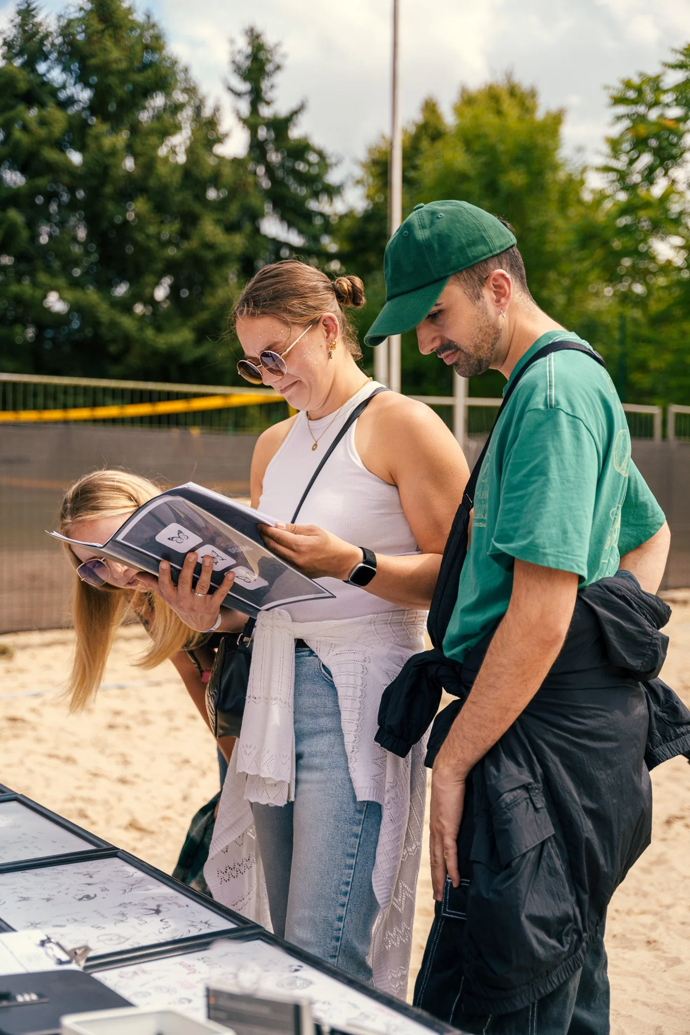 Three people looking at drawings or artwork displayed on a table outdoors on a sunny day, with trees in the background.
