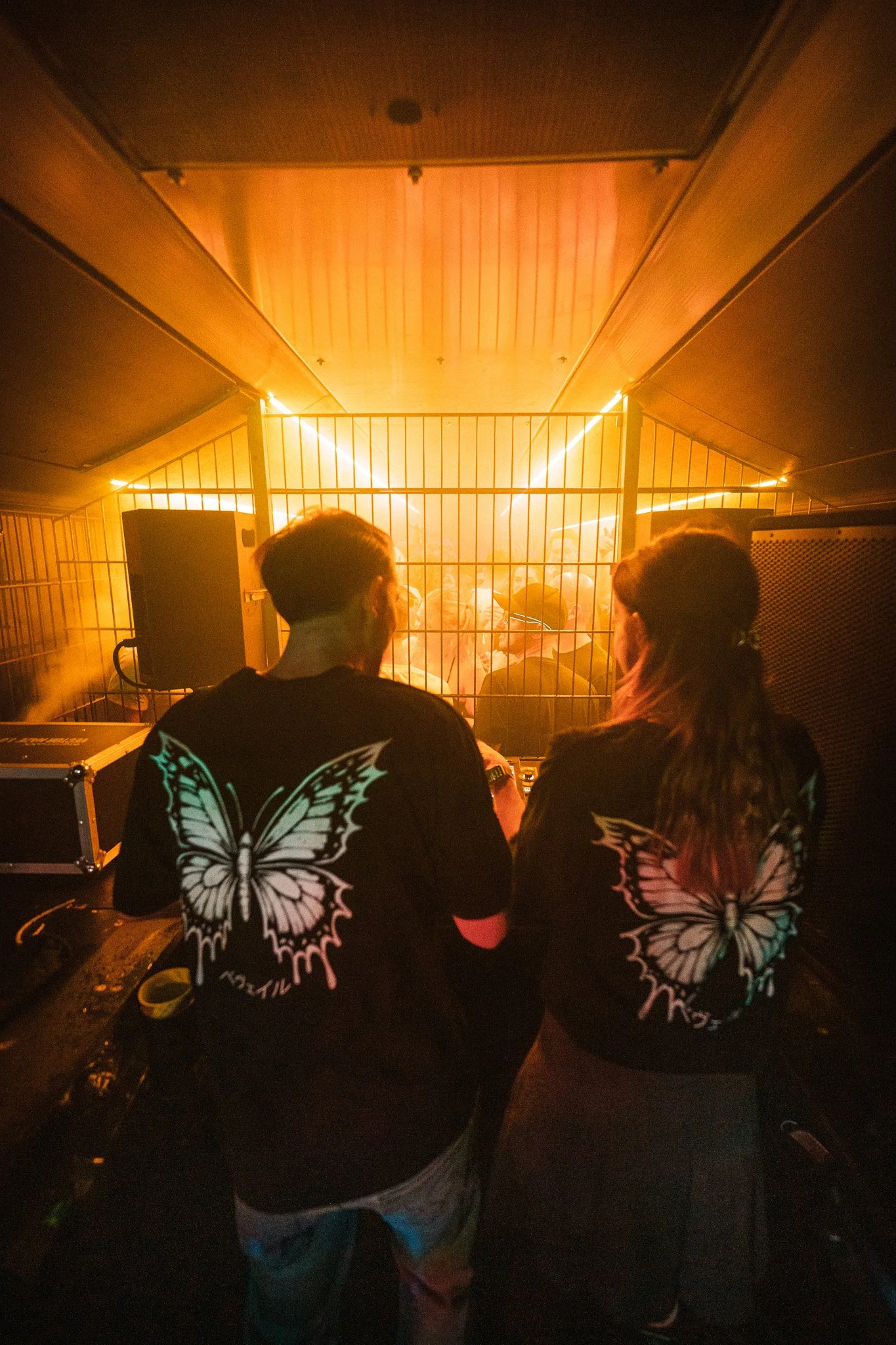 Two people standing behind a DJ booth at a nightclub with a crowd dancing in the background, both wearing black shirts with large butterfly designs on the back.