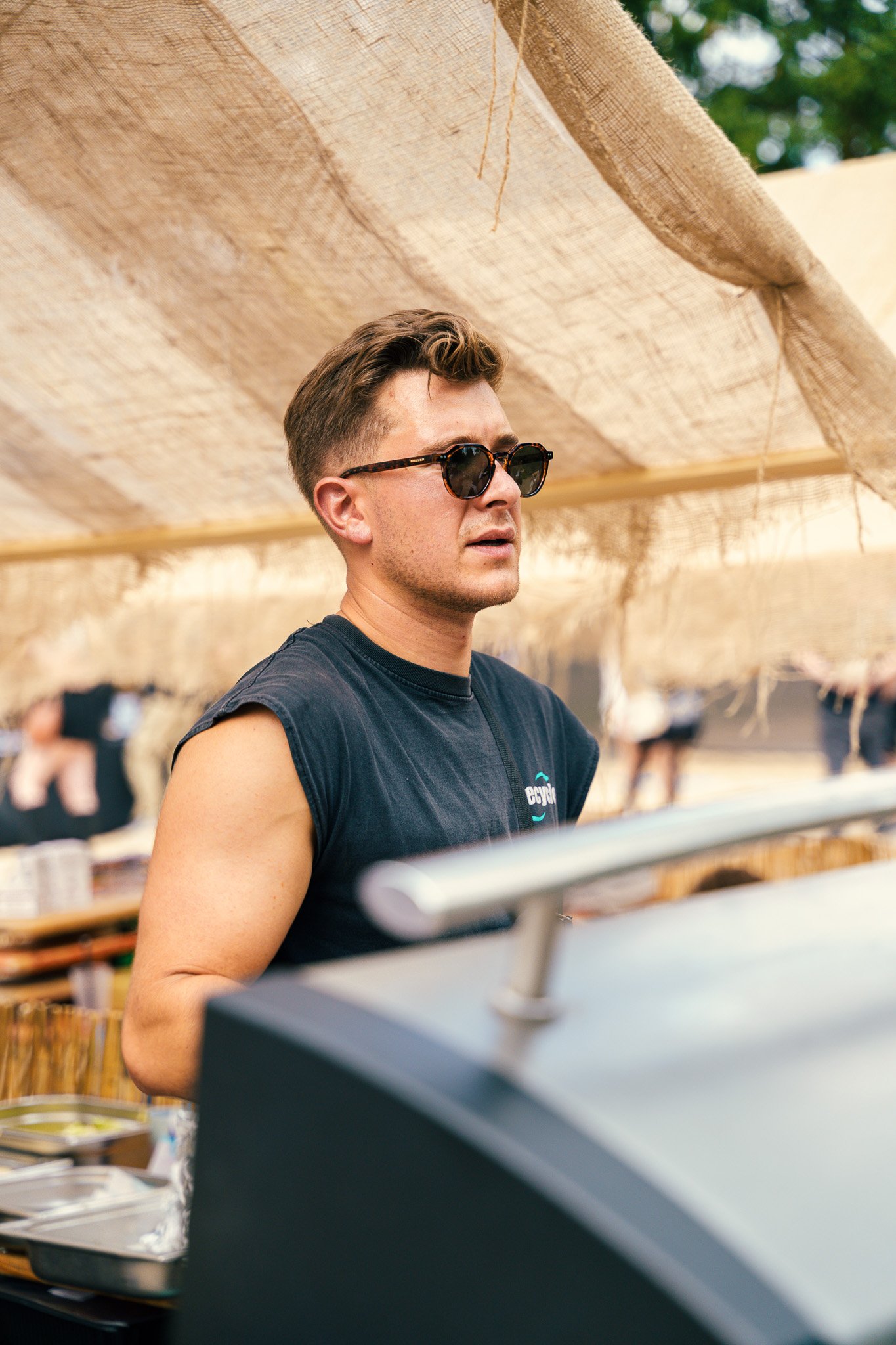 Young man with short hair and sunglasses at outdoor event, standing behind a grill or serving station, with a beige canopy overhead and people in the background.