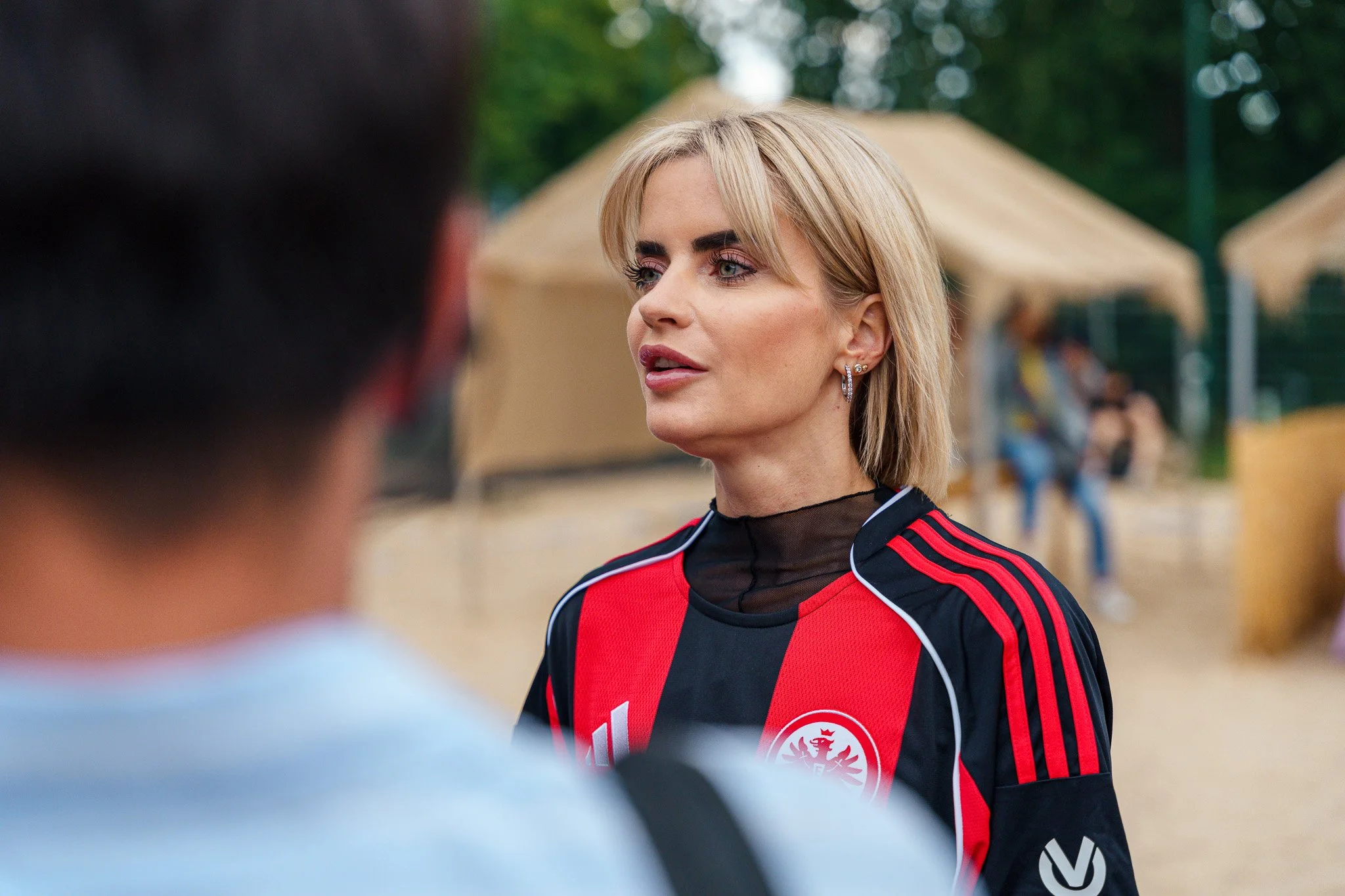 A woman with blonde hair wearing a black and red sports jersey and earrings, speaking to a person whose back is visible, outdoors with tents and people in the background.