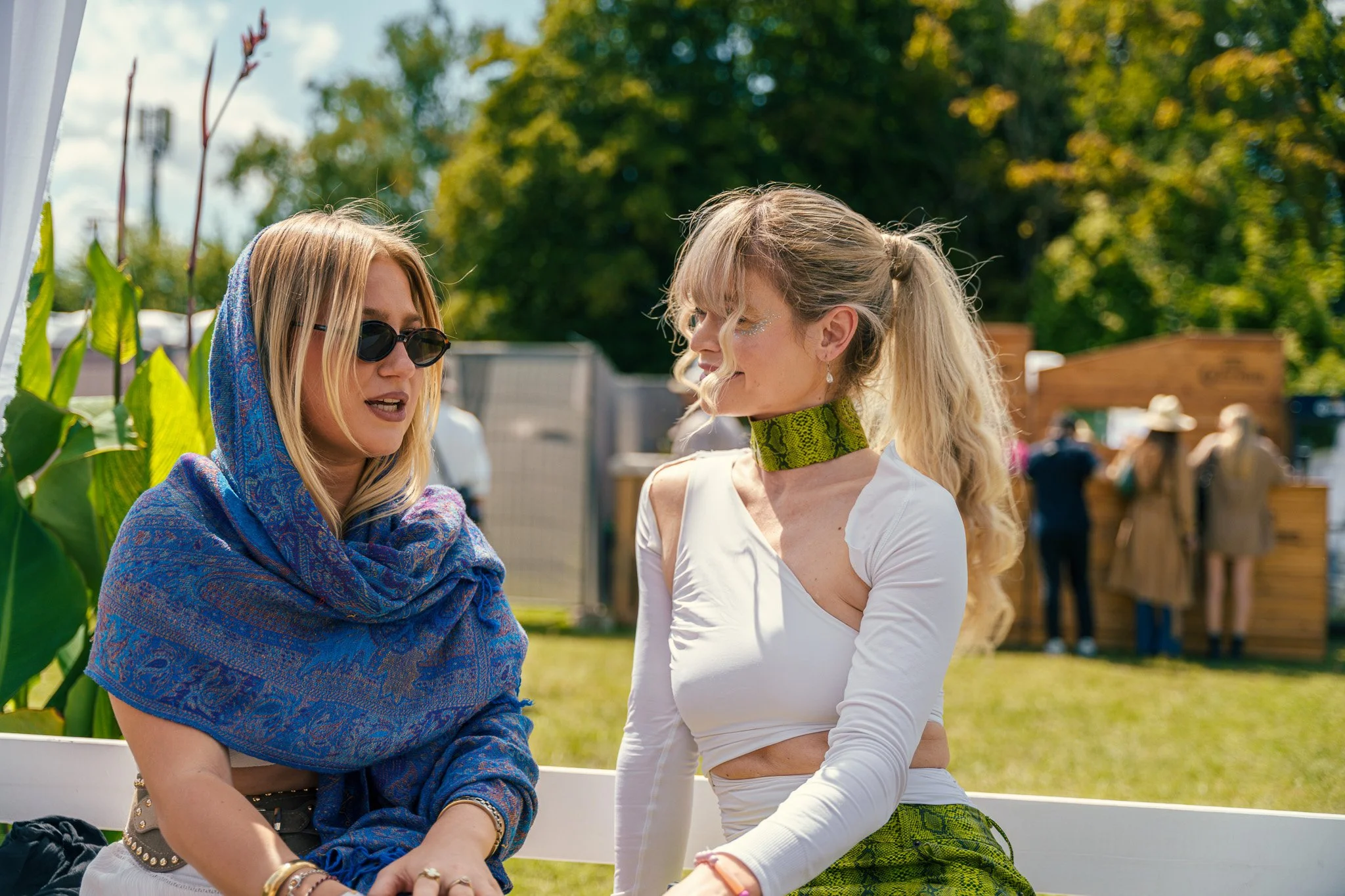 Two women sitting on a white bench at an outdoor event, engaging in conversation. One woman is wearing sunglasses, a blue patterned scarf, and a beige top. The other woman is dressed in a white crop top with cut-out shoulders, green snake print skirt