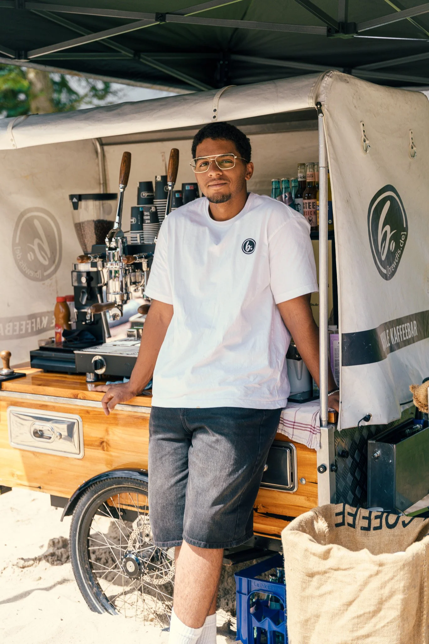 A man standing behind a mobile coffee stand with espresso machine, wearing glasses, white T-shirt, and shorts, outdoors on a sunny day.