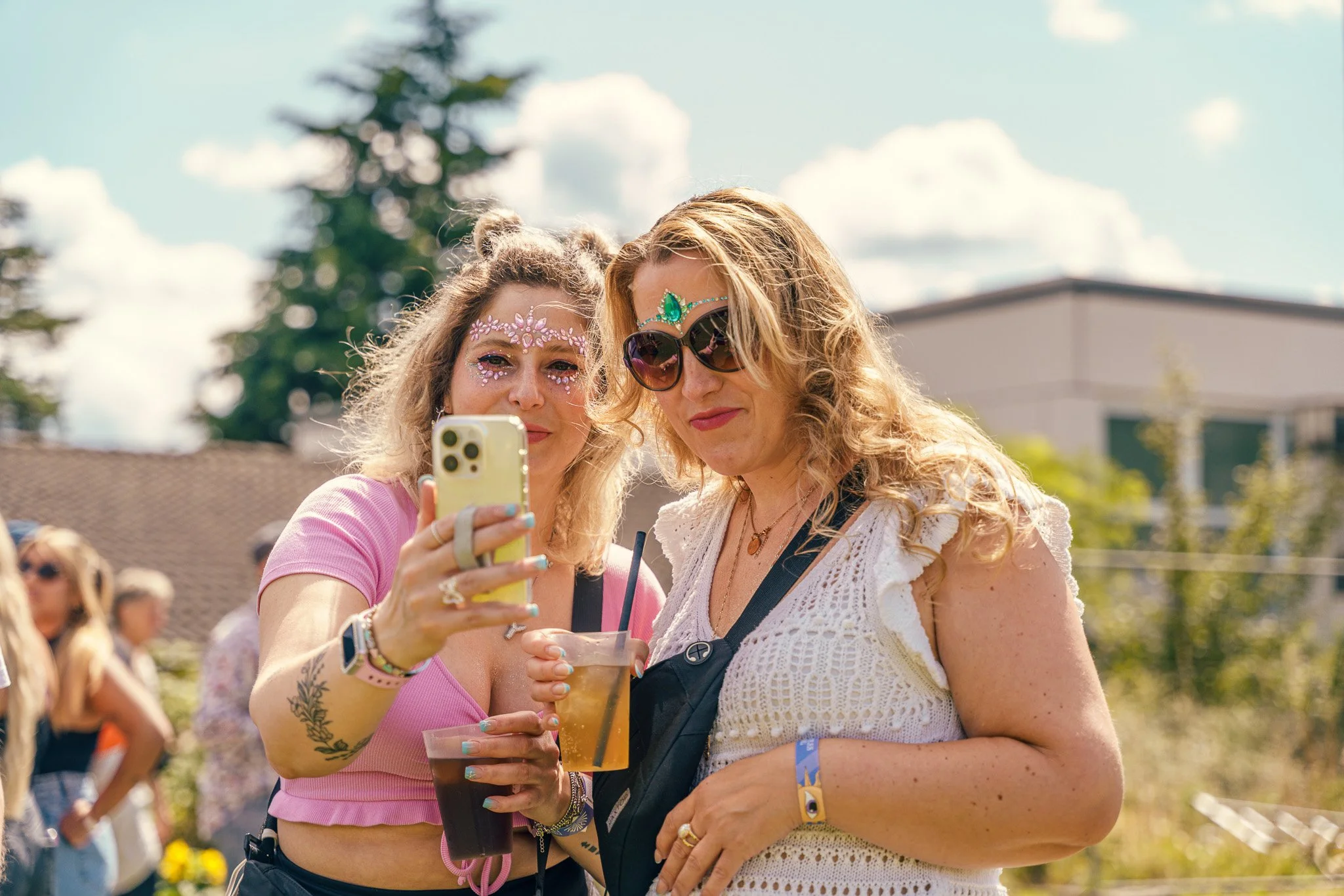 Two women taking a selfie at an outdoor event, one with face glitter and the other with sunglasses and face jewelry, holding drinks.