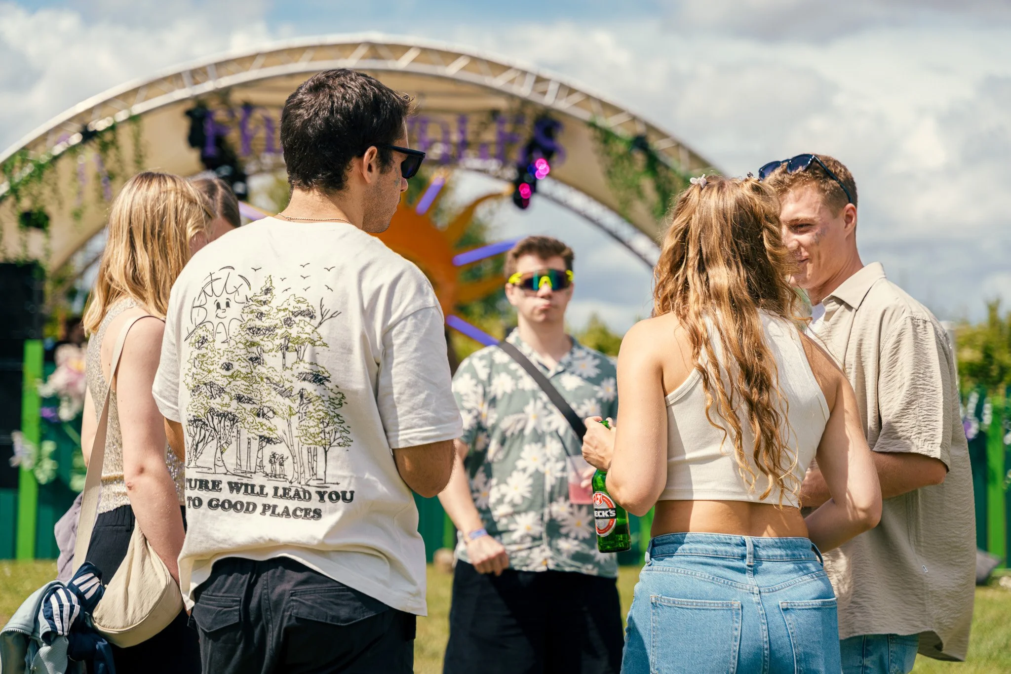 Group of young people at an outdoor music festival, with a stage in the background, enjoying a sunny day, some wearing sunglasses, one holding a beer bottle.