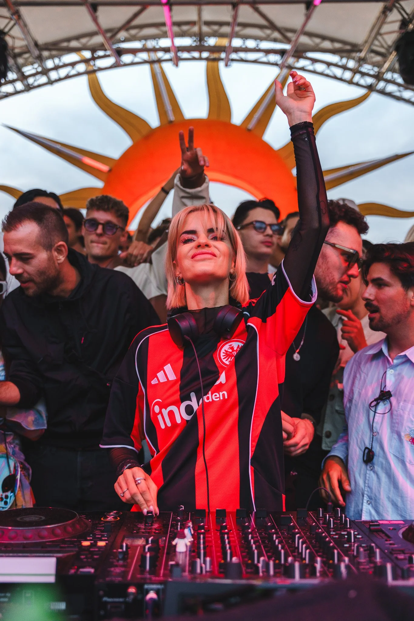A female DJ cheering at a music event with a crowd behind her, in front of a large sun-shaped stage backdrop.