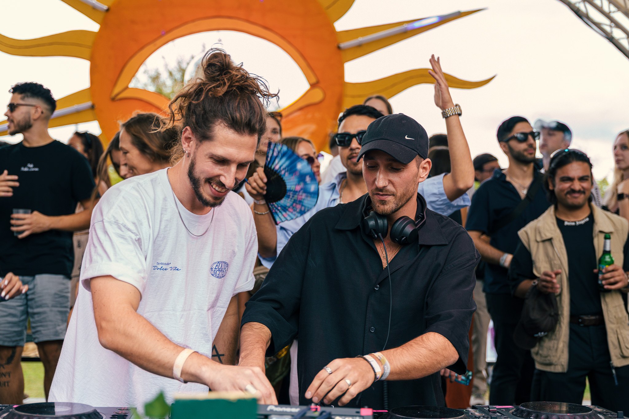 Two DJs perform at an outdoor event with a crowd dancing and watching in the background, and a large yellow and orange decorative structure behind them.