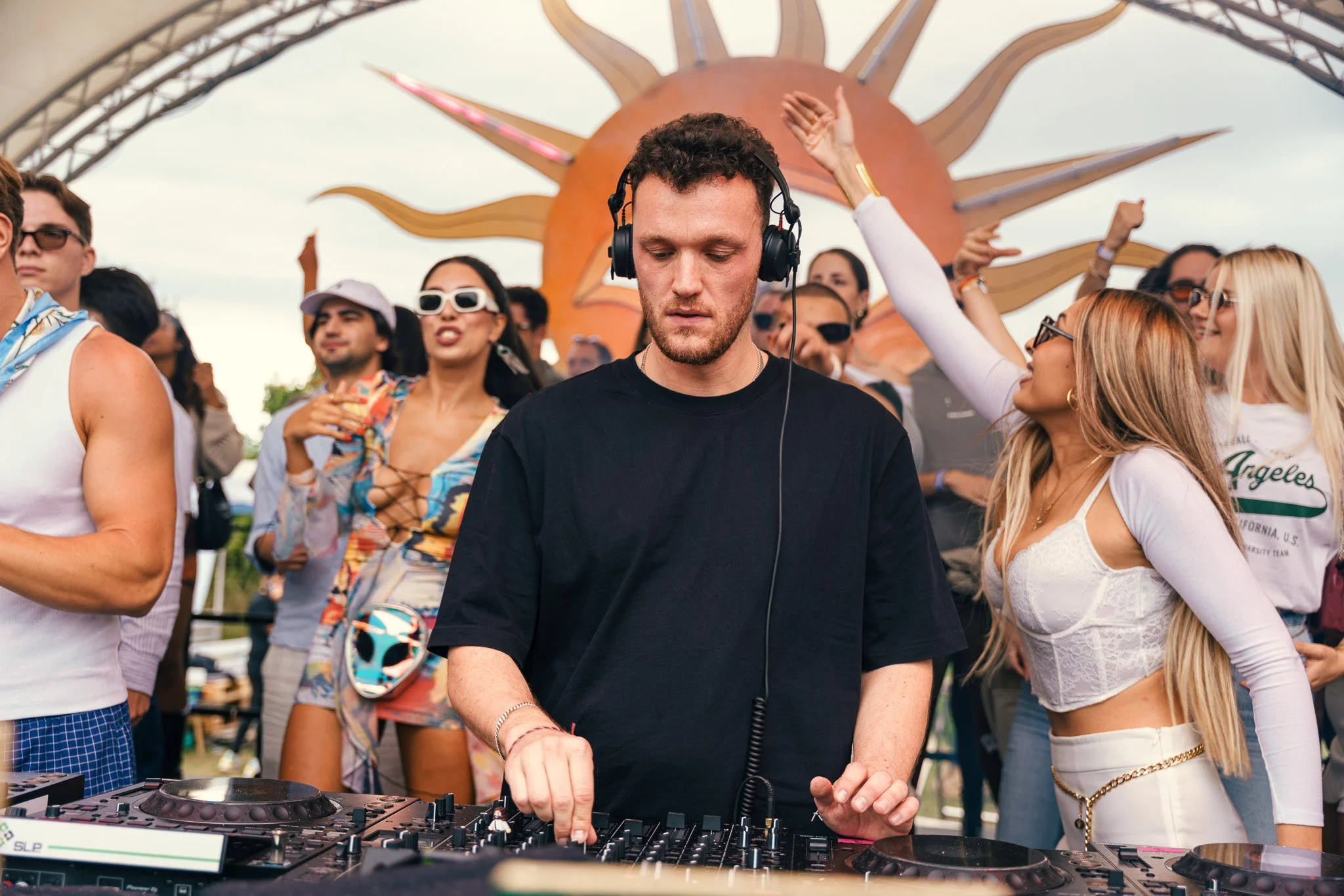 A DJ in black shirt wearing headphones plays music at an outdoor festival with a crowd dancing and enjoying in the background.