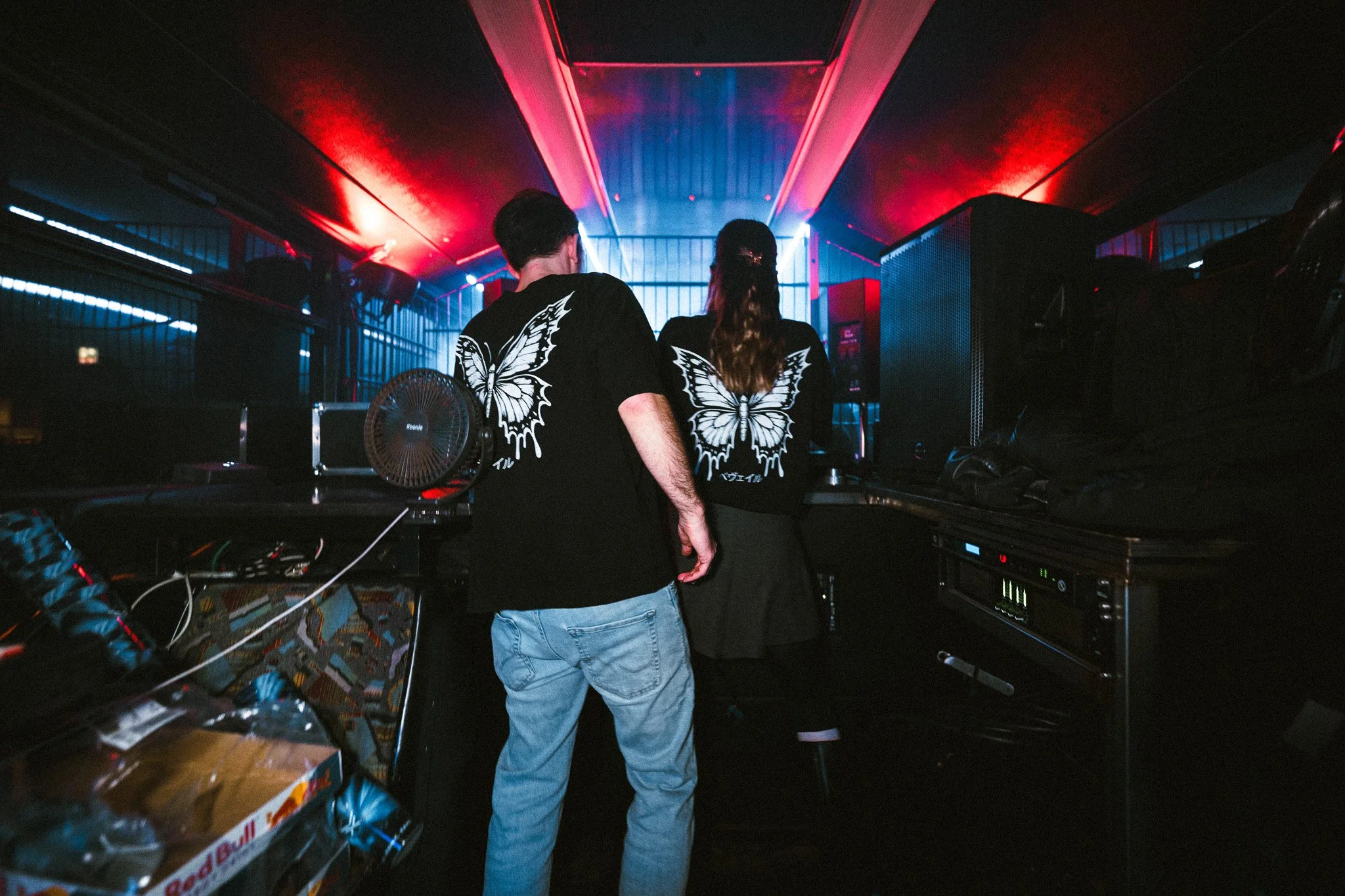 Two people standing inside a dimly lit nightclub or event space with blue and red lighting, wearing matching black shirts with large butterfly designs on the back, facing a stage or DJ area.
