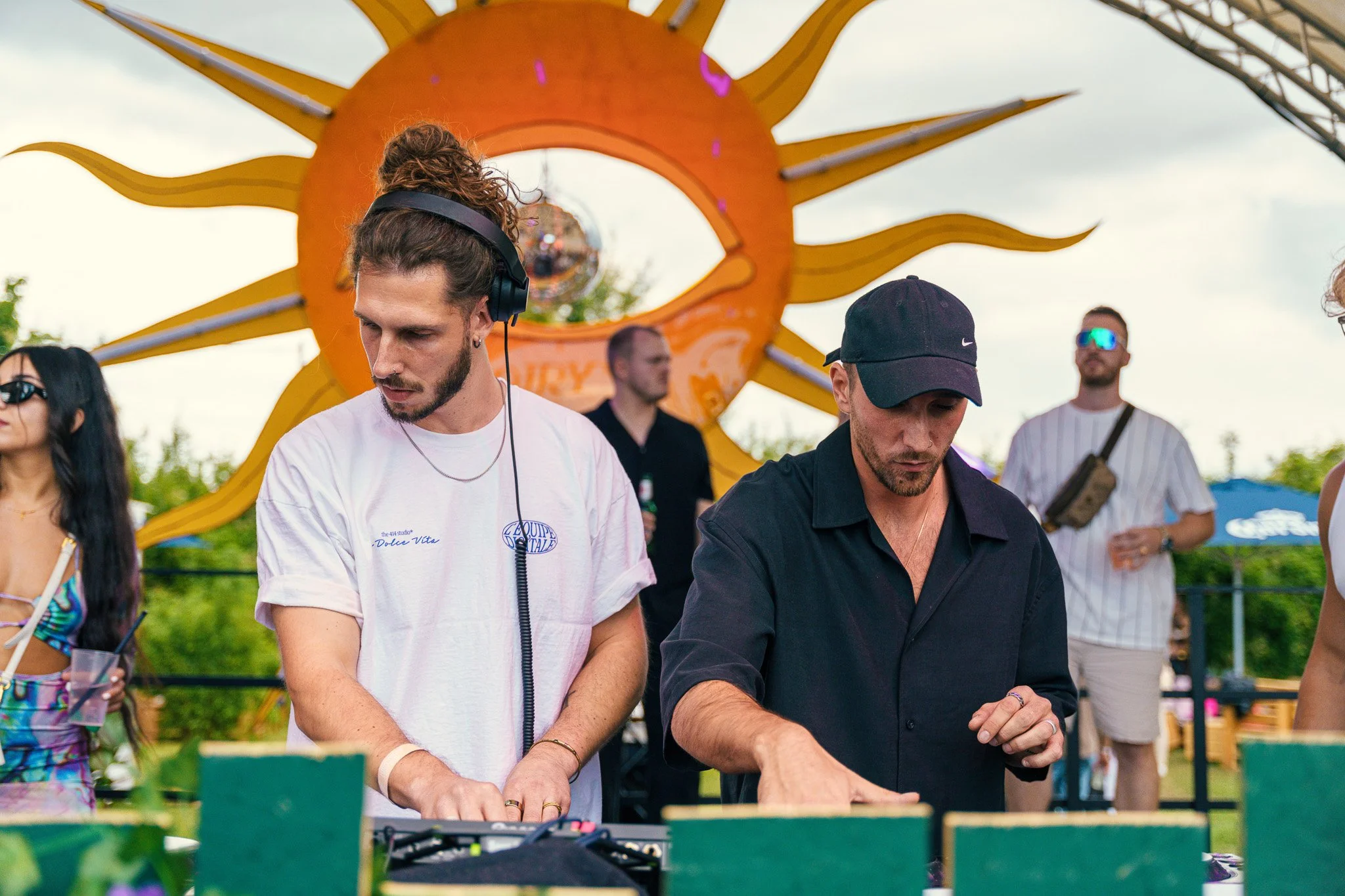 Two male DJs playing music at an outdoor event, with a large decorative yellow sun sculpture in the background and other people standing nearby.
