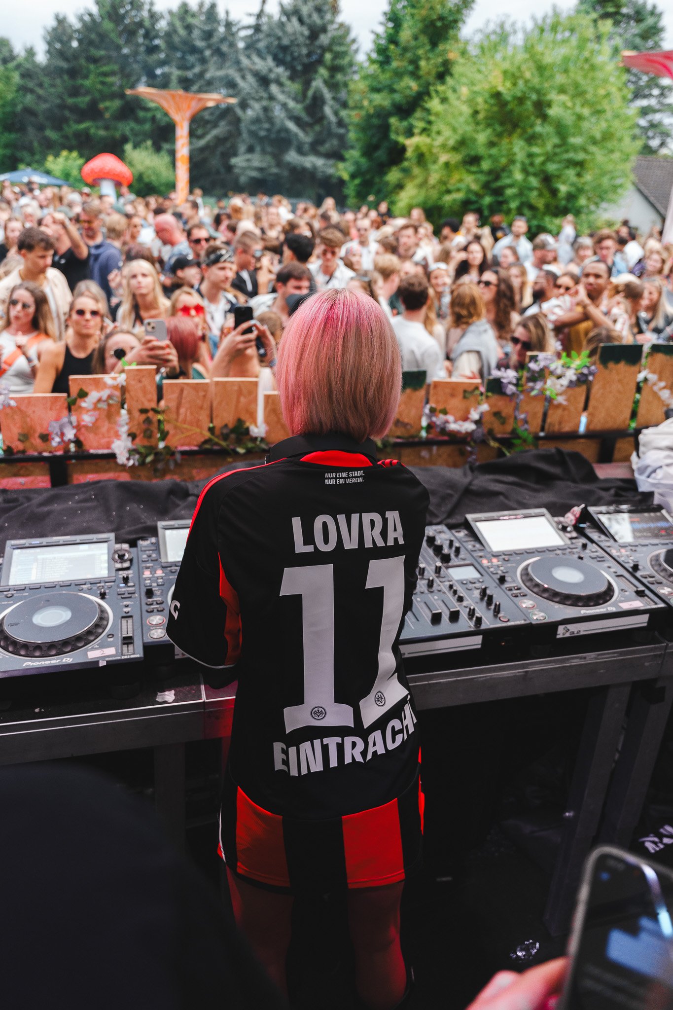 A DJ with pink hair standing behind DJ equipment at an outdoor music event, facing a large crowd of people gathered in front of a stage, with trees and clouds in the background.