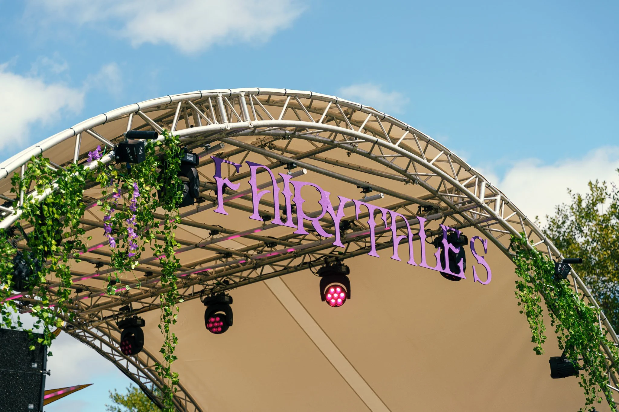 Concert stage arch with purple lettering reading 'FLOWERFEST', decorated with hanging green vines and purple flowers, under a clear blue sky.