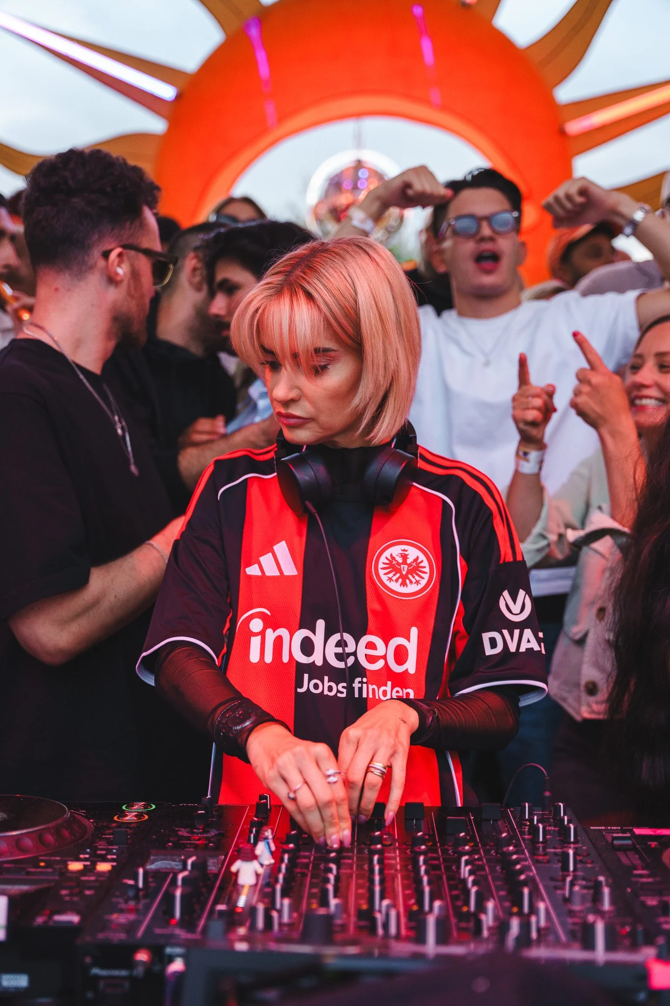 Female DJ with blonde hair and headphones around her neck, wearing a red and black sports jersey, mixing music at a party with a crowd of people dancing and cheering behind her.