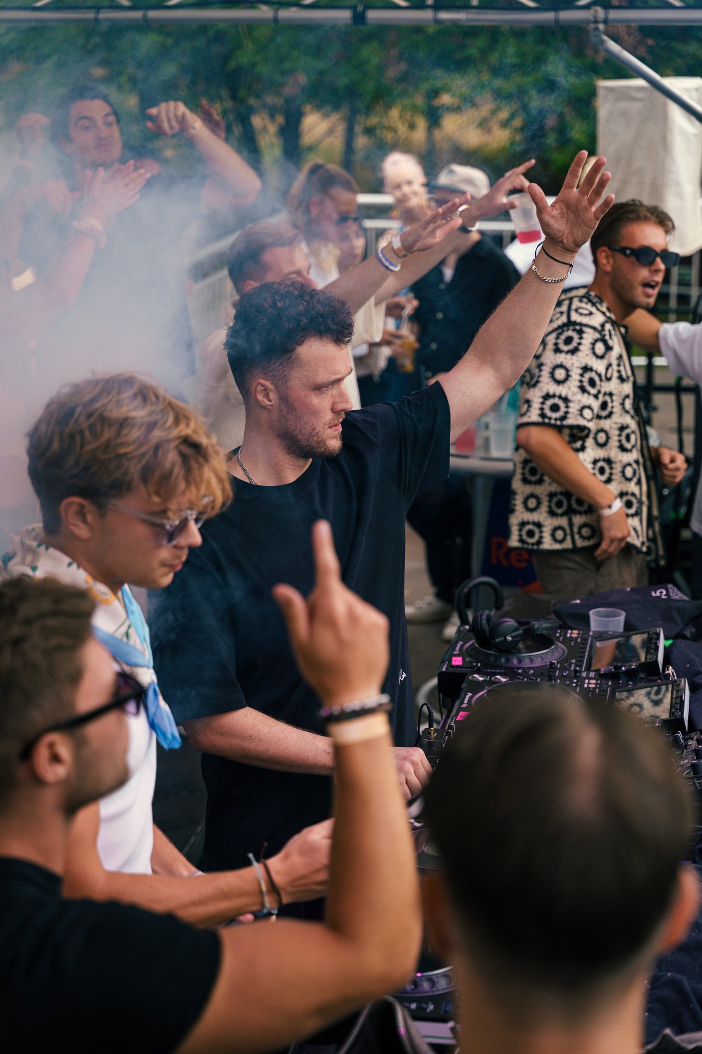 A DJ performs at an outdoor music event, raising one hand in front of a crowd dancing and enjoying the music.