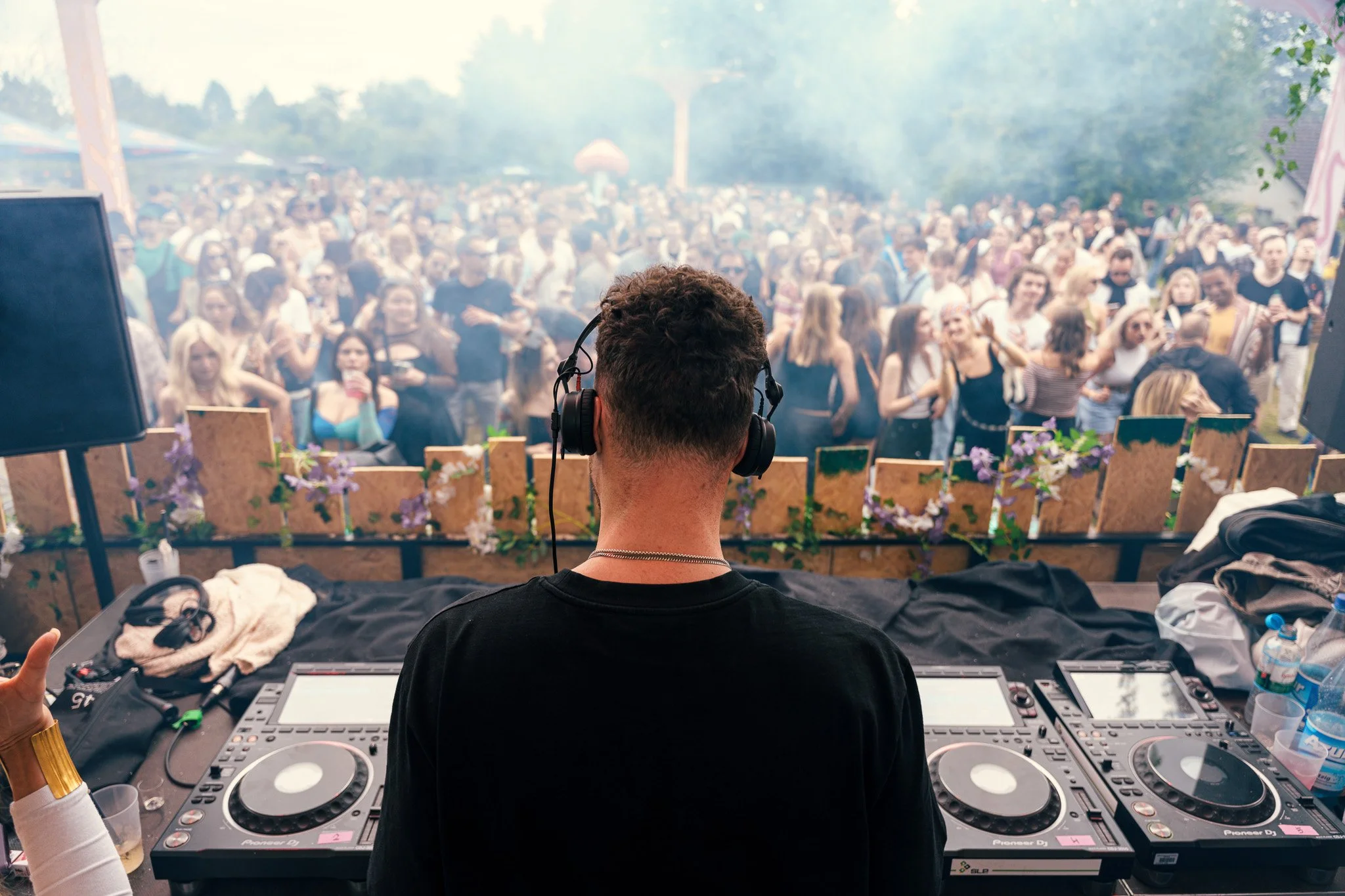 DJ performing at an outdoor music festival, facing a large crowd of people dancing and enjoying the event with mist or smoke in the air.