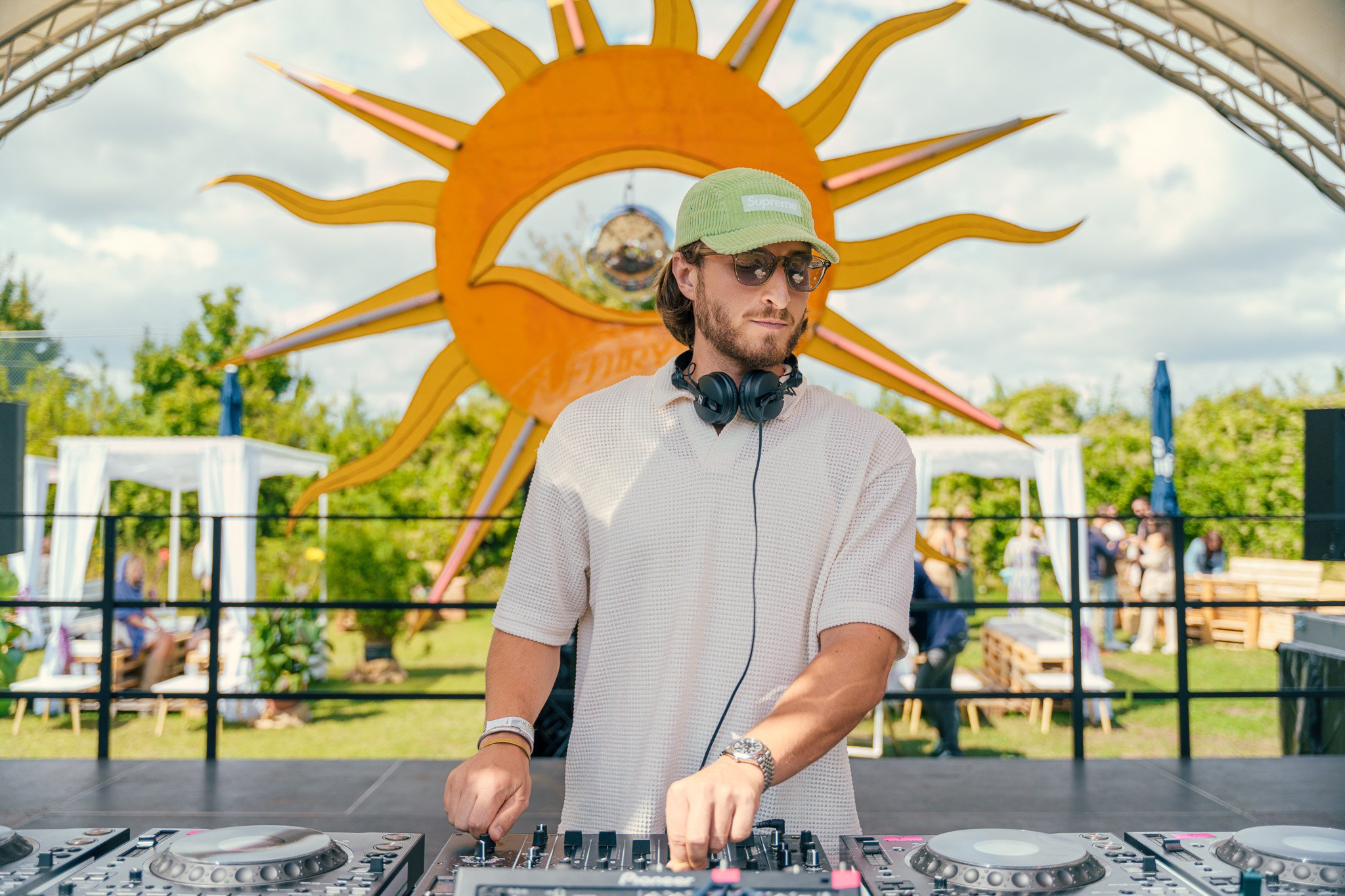 A male DJ wearing sunglasses, a green cap, and headphones around his neck, standing behind DJ equipment outdoors during daytime with a decorative sun and eye art installation behind him.