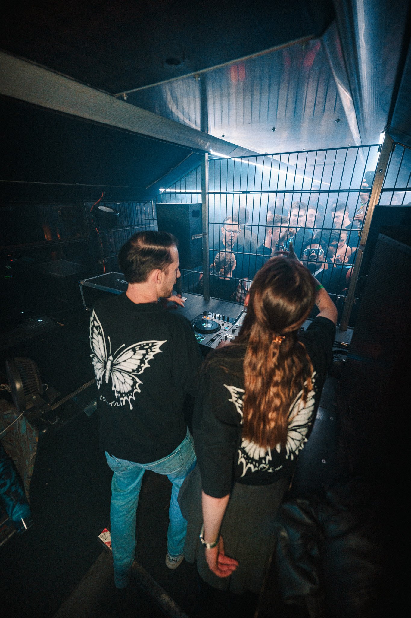DJ playing music at a nightclub with a crowd dancing behind a metal fence.