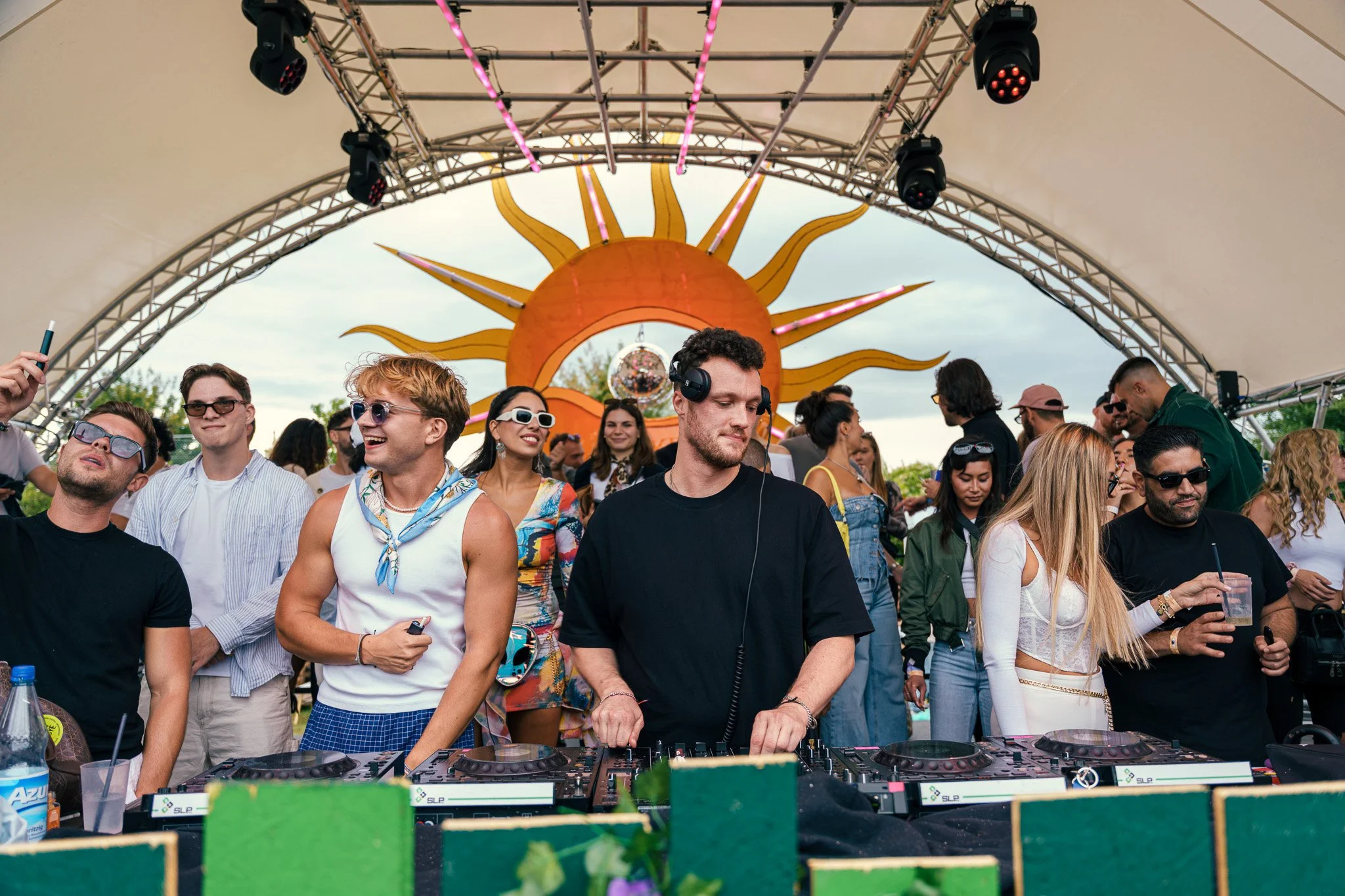 A DJ with headphones mixes music at an outdoor event under a canopy with a sun decoration in the background, surrounded by a diverse crowd dancing and enjoying the music.