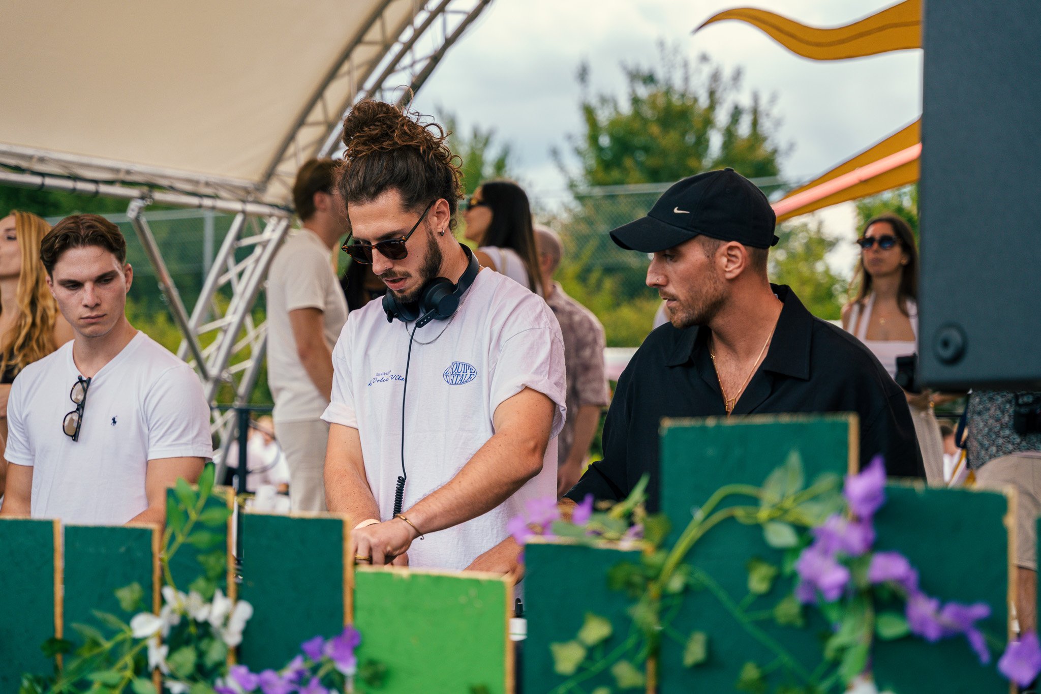 A DJ playing music at an outdoor event, surrounded by people and decorated with purple flowers and colorful panels.