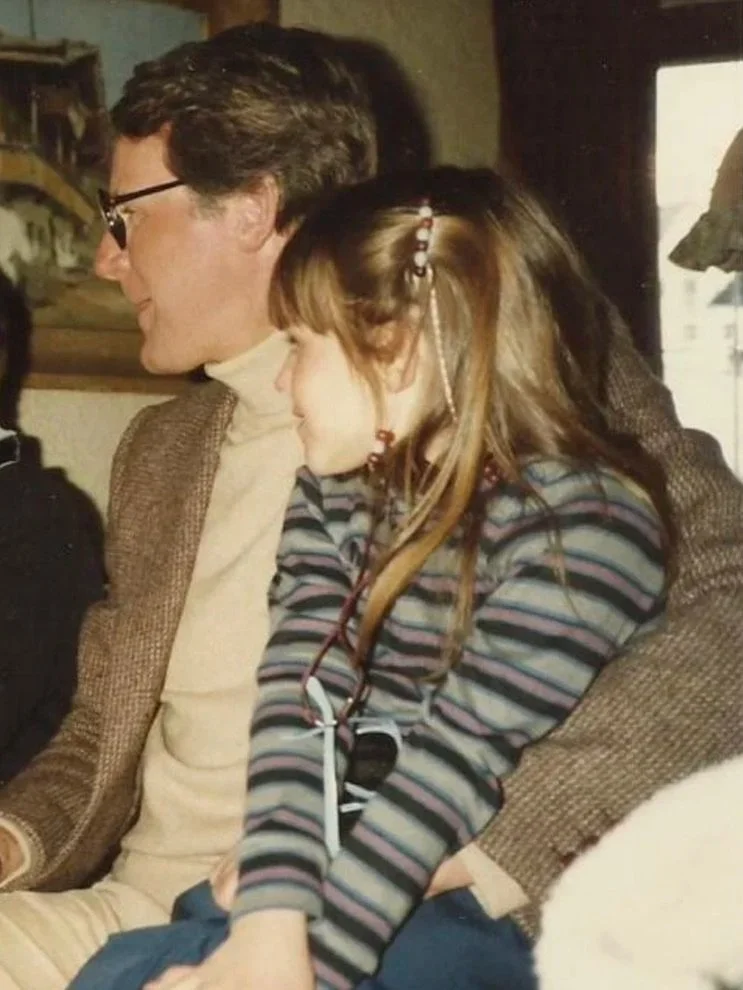 A man and a young girl sitting closely together, with the girl resting her head on the man's shoulder in an indoor setting.