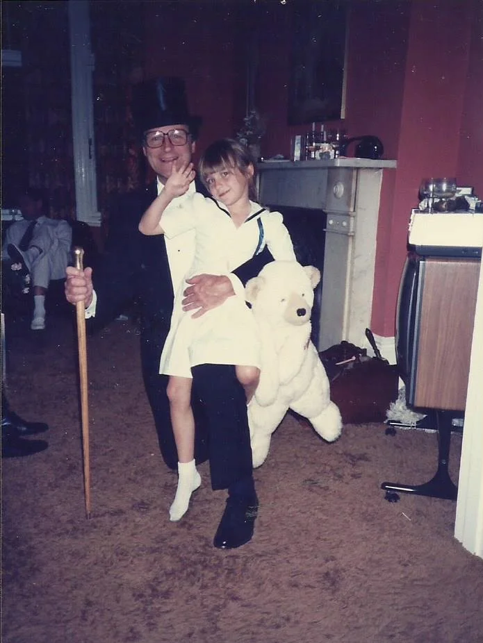 A man in a tuxedo with a top hat, glasses, and a walking cane is holding a young girl dressed in a white dress. They are sitting on a giant plush polar bear in a room with a fireplace, a wooden cabinet, and other furniture, likely in a home.