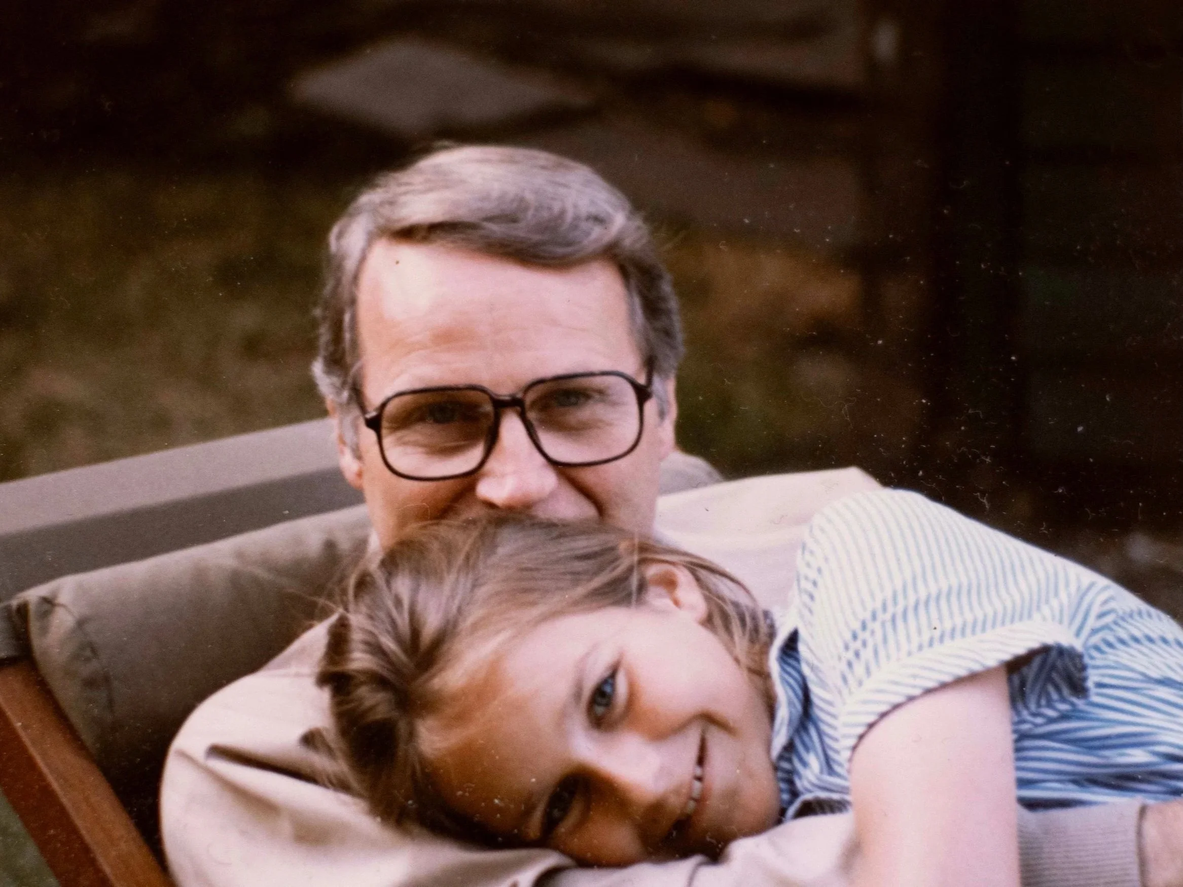 A man with gray hair and glasses embracing a young woman with brown hair and a striped shirt, both lying on a sofa.
