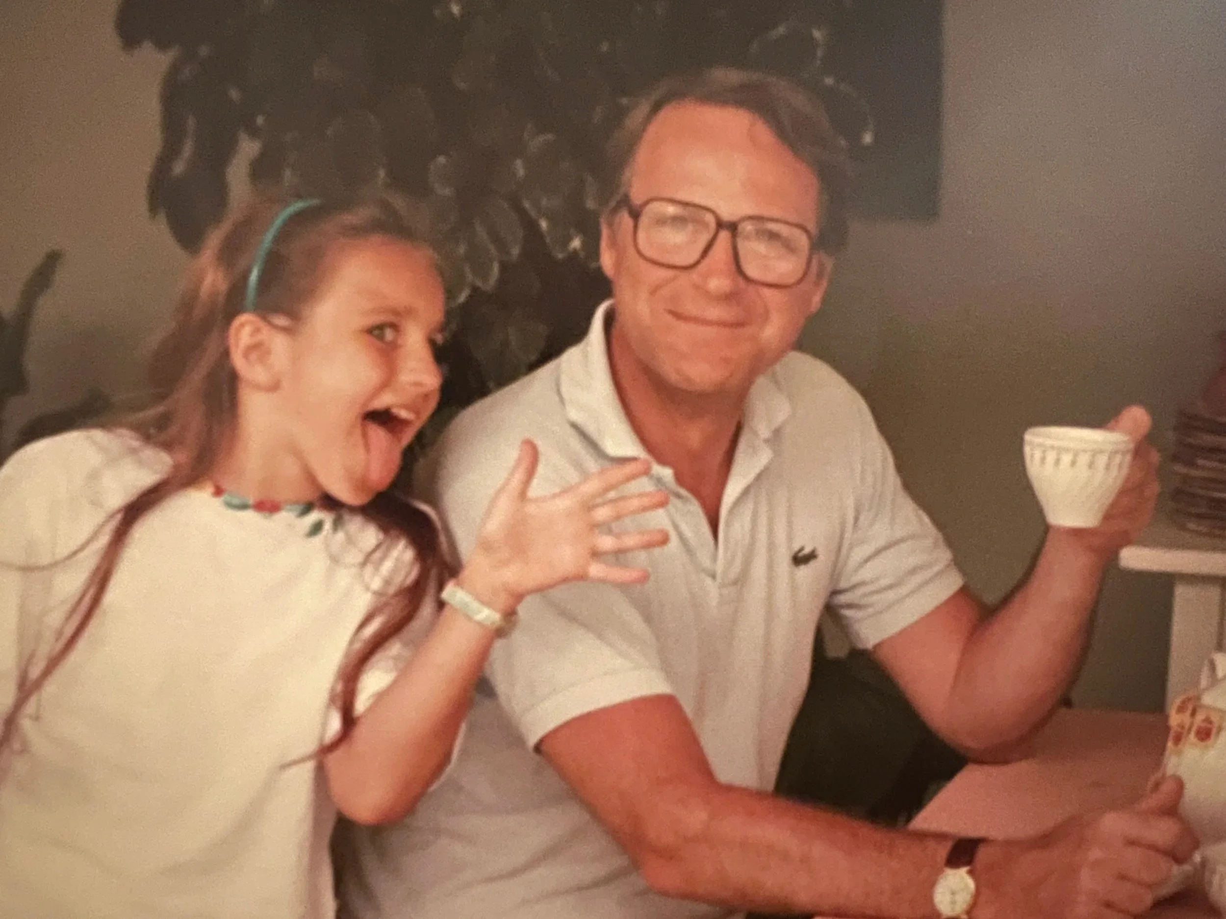 A young girl and an older man smiling and sitting at a table, the girl appears to be excited or talking, holding her hand up. The man wears glasses, a white shirt, and holds a cup in his hand. The background is dark with a patterned wall.
