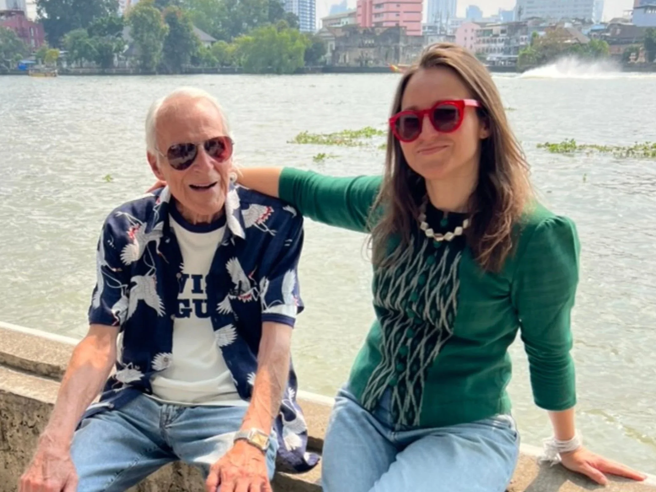 Two women sitting by the water, smiling. One woman has white hair, wearing sunglasses, a floral shirt, and a white T-shirt. The other woman has brown hair, red sunglasses, and a green jacket. Buildings are in the background across the water.