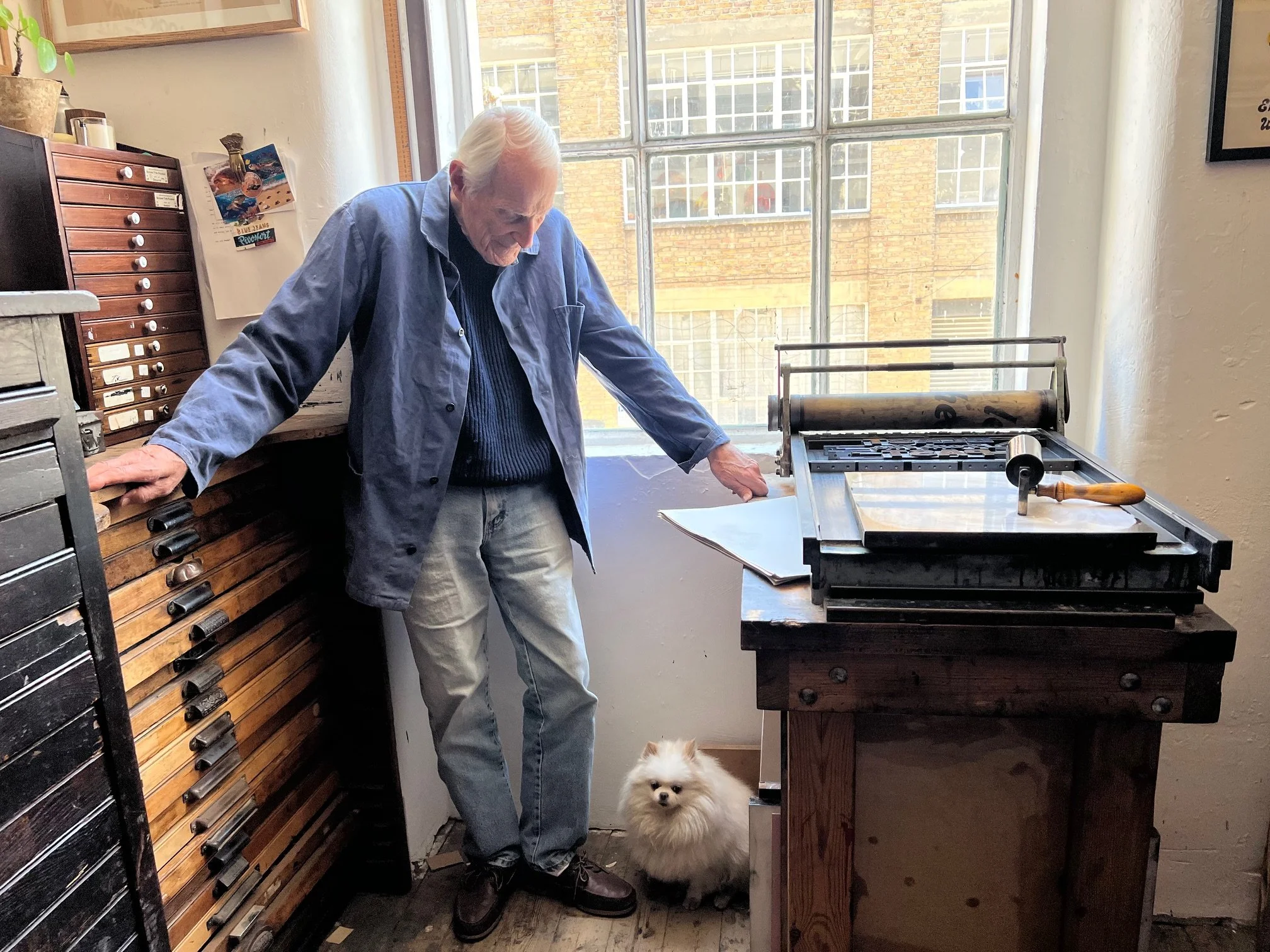 An elderly man with gray hair leaning on a kitchen counter, looking down. A small white dog with fluffy fur sitting on the floor looking forward. A large window with a brick building outside, a wooden cabinets and a rolling pin on a vintage pasta mac