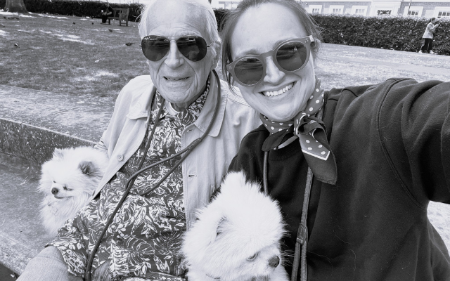 A young woman and an elderly man sitting on a park bench, smiling for a selfie, each holding a small fluffy white dog. They are wearing sunglasses, and the woman has a polka dot scarf, in an outdoor setting with a grassy area and buildings in the background.