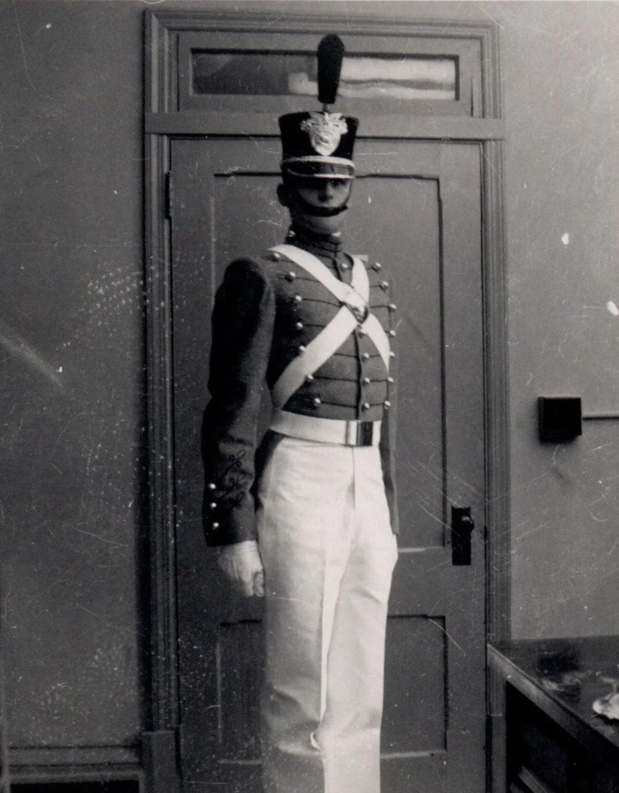 Black and white photo of a soldier in a formal military uniform standing in front of a door. The soldier is wearing a tall hat with a decorative emblem, a dark jacket with buttons, sash, and light-colored trousers.