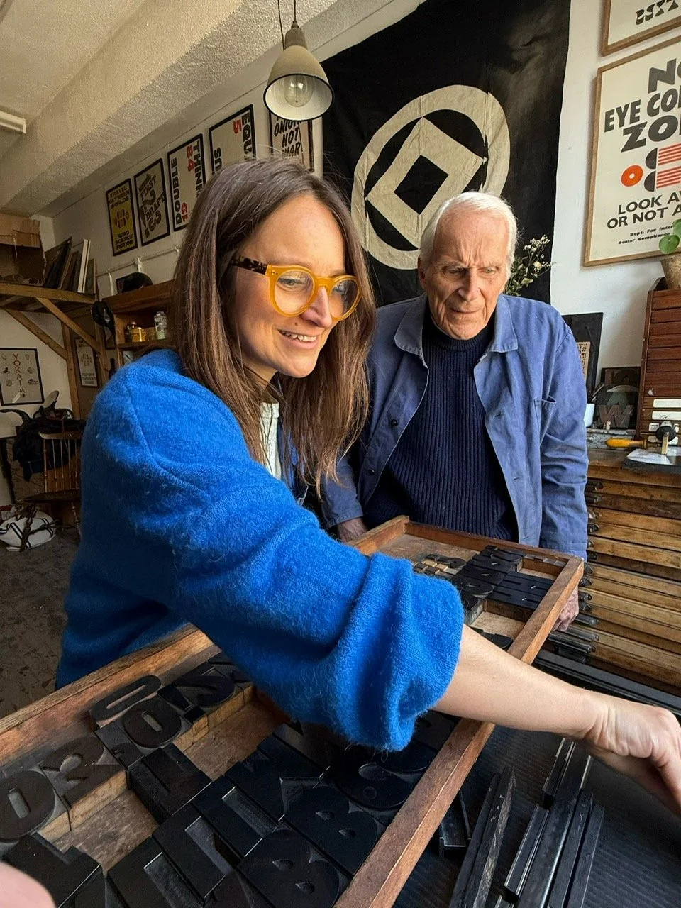 A woman with yellow glasses and a blue sweater is arranging black metal letterpress blocks on a wooden surface. An elderly man stands behind her, observing. The background includes framed posters, a black flag with a white symbol, and various art sup