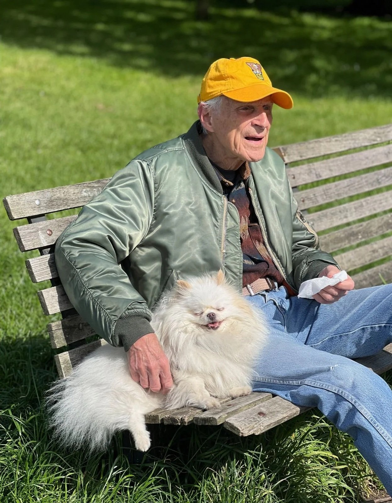 An elderly man with a yellow cap, wearing a silver jacket, and blue jeans sitting on a wooden park bench outdoors, petting a small fluffy white dog that appears to be relaxed and happy.