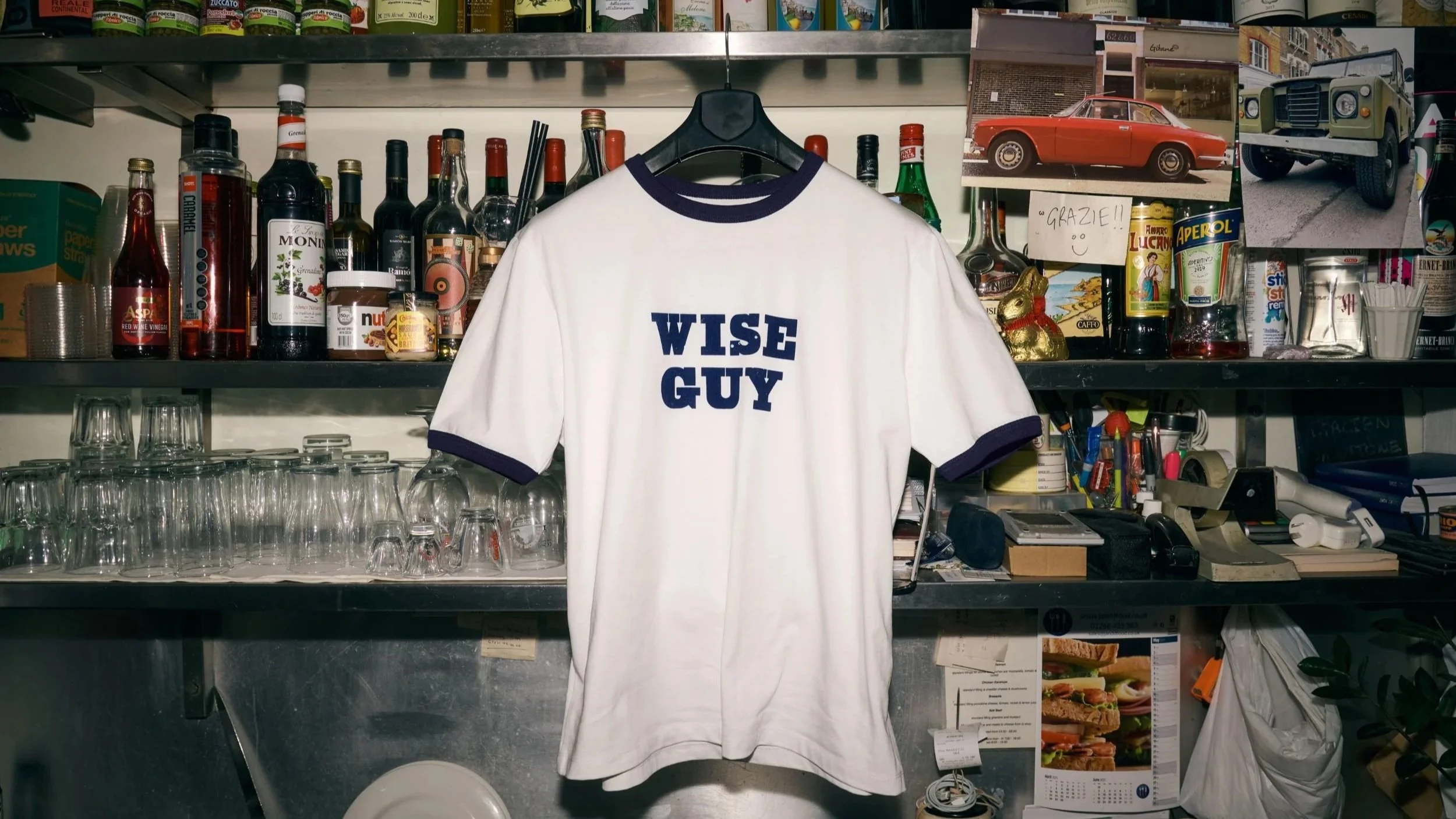 A white baseball jersey with navy blue trim and the words "WISE GUY" printed on the front, hanging on a black hanger against a cluttered kitchen shelf with bottles, glasses, and photos.