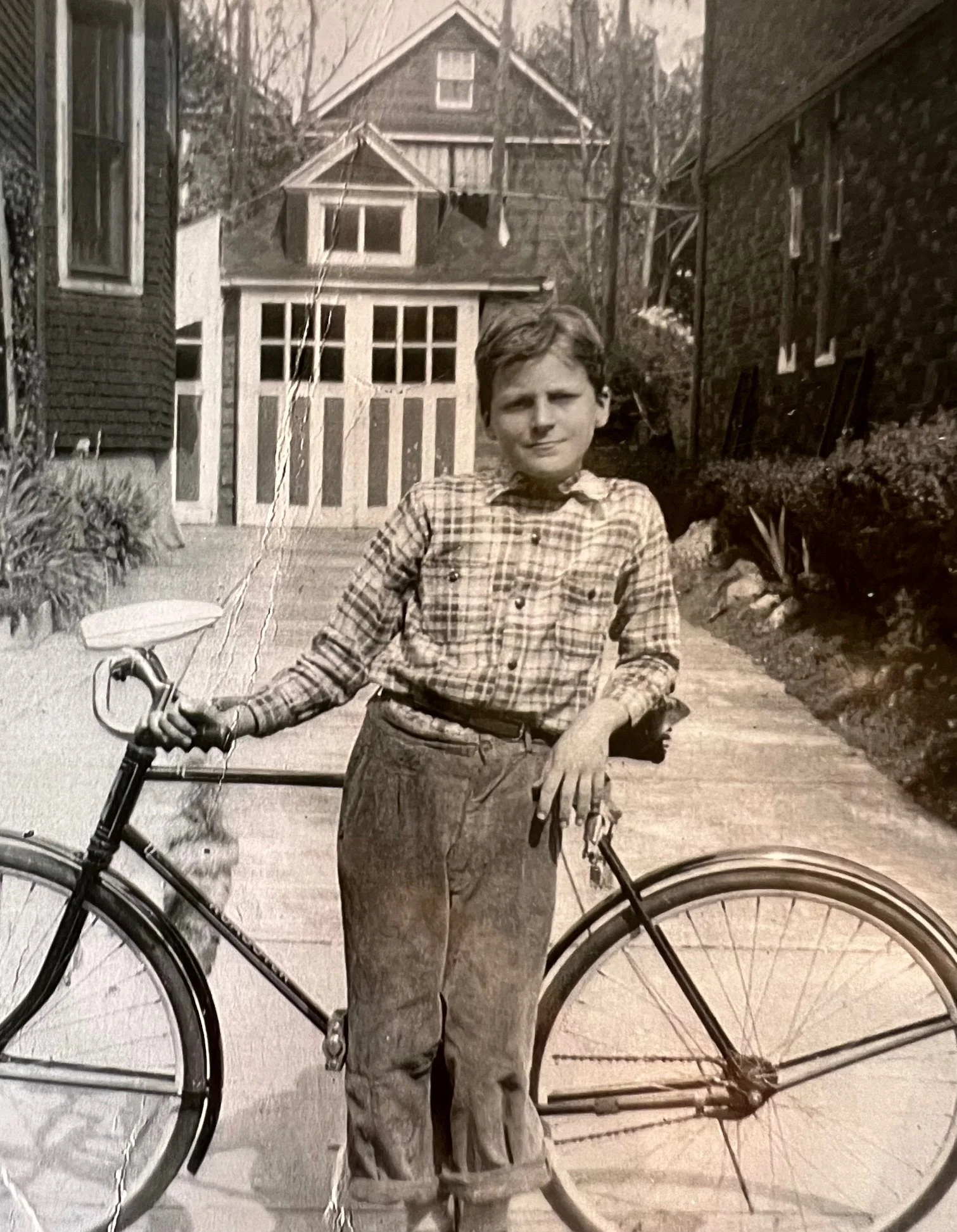 A young boy standing next to a bicycle in front of a house, in black and white.