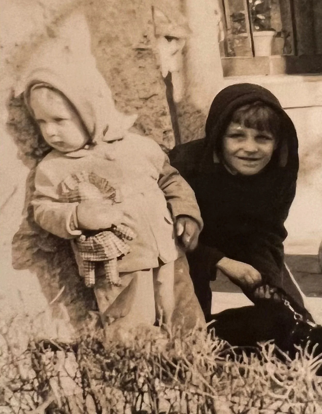 A young girl with a hood and coat standing next to a woman with a head covering and dark clothing, both kneeling outside near a rock formation, with rocks and grass in the foreground and a small shelf with containers in the background.