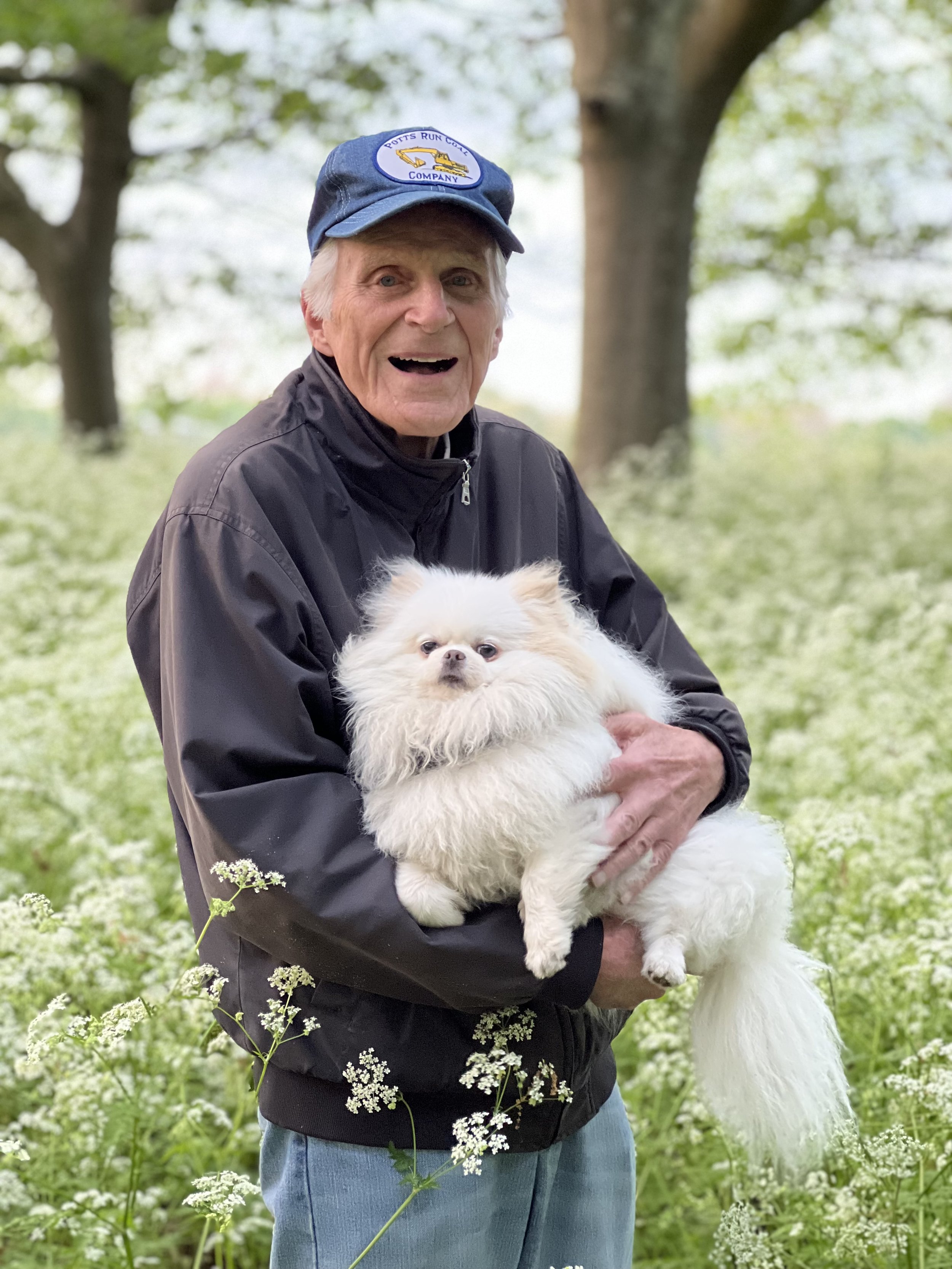 An elderly man wearing a blue cap and black jacket holding a fluffy white dog outdoors in a field of white flowers with trees in the background.