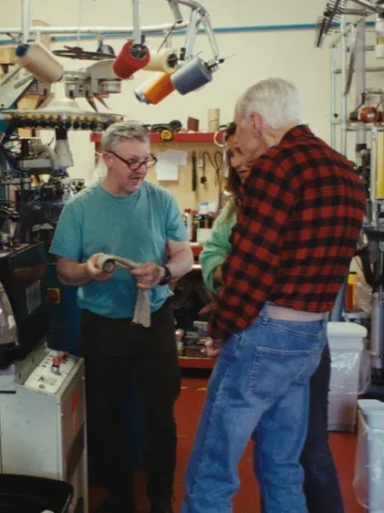Two men and a woman in a workshop surrounded by tools and equipment, engaged in a conversation. The woman is holding a wooden object, and both men are attentively listening.