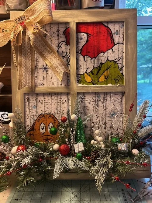 A holiday decor display with a rustic wooden window frame featuring a Santa hat, and Christmas ornaments and greenery with a cartoon cat face and a character with a red Santa hat and yellow boots in the background.