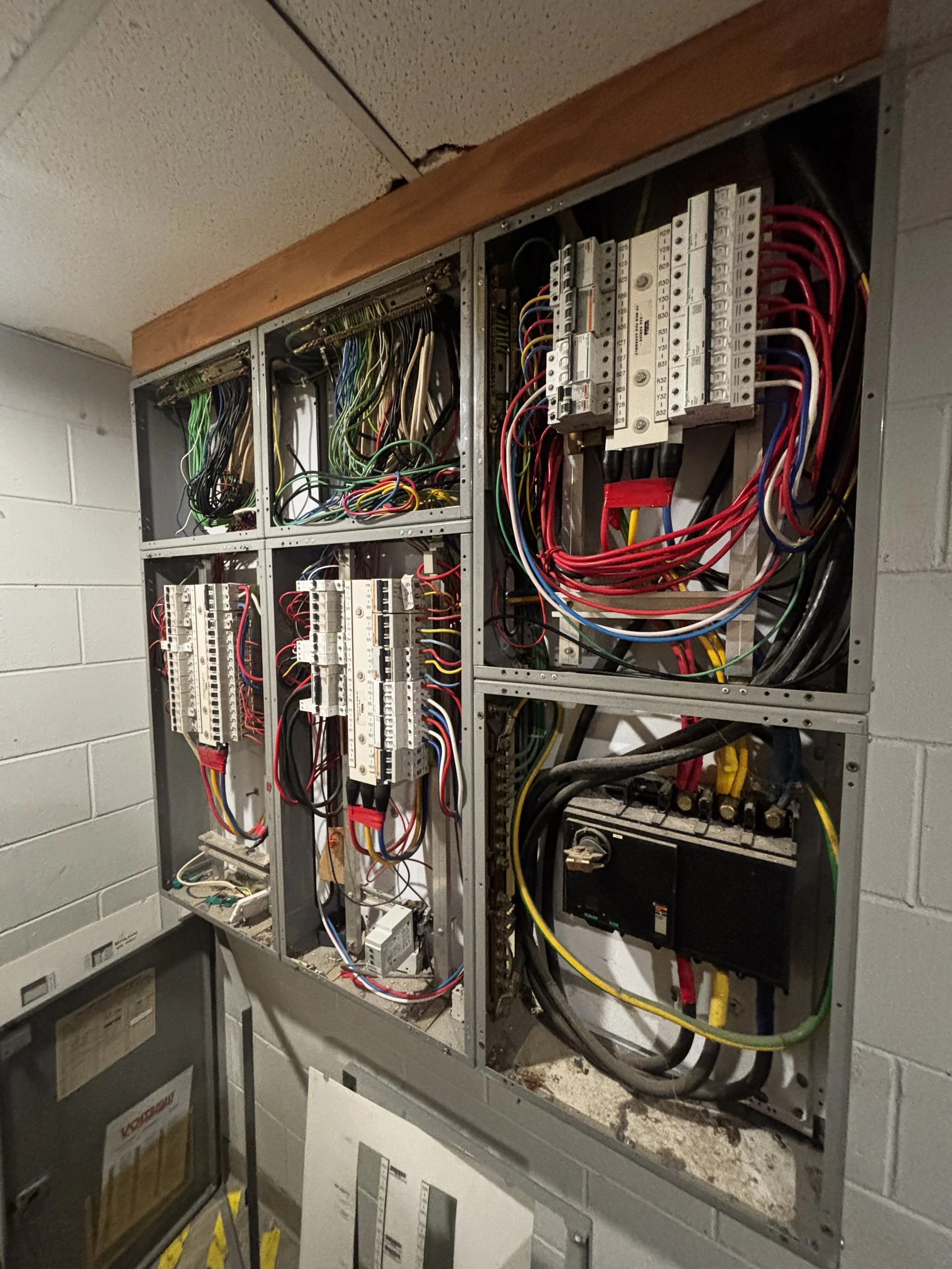 An electrical panel with multiple circuit breakers and wiring inside an open metal box, mounted on a brick wall.