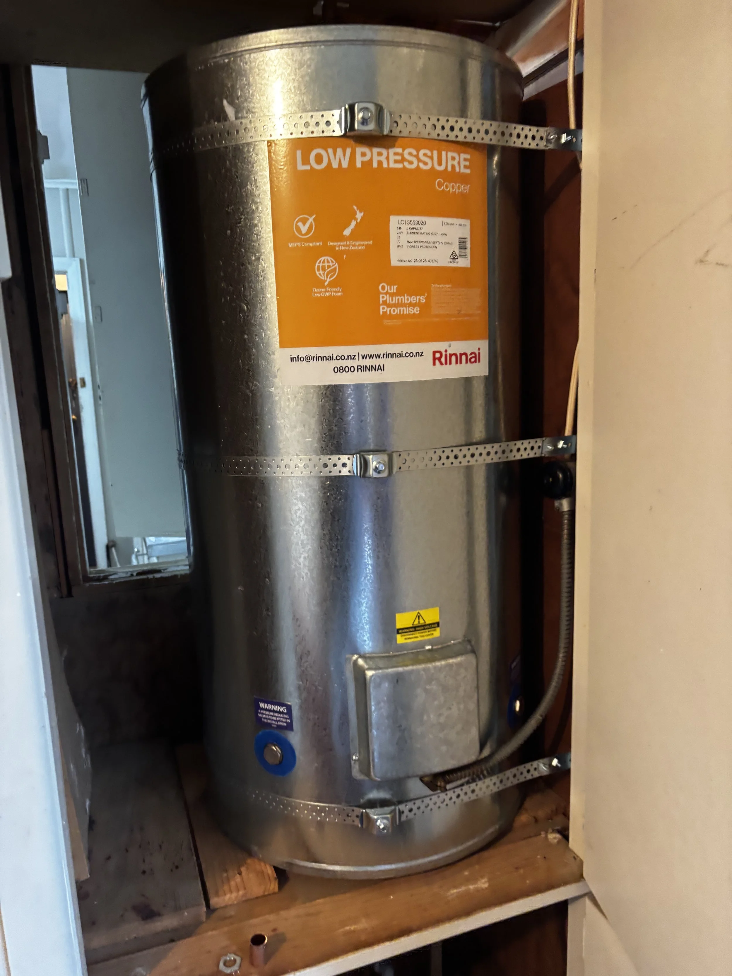 A large, cylindrical water heater with a silver metallic finish, mounted in a wooden cabinet. The heater has a yellow label indicating low pressure, with additional labels and warning stickers. There is a visible pipe connected at the bottom and a power cord on the side.