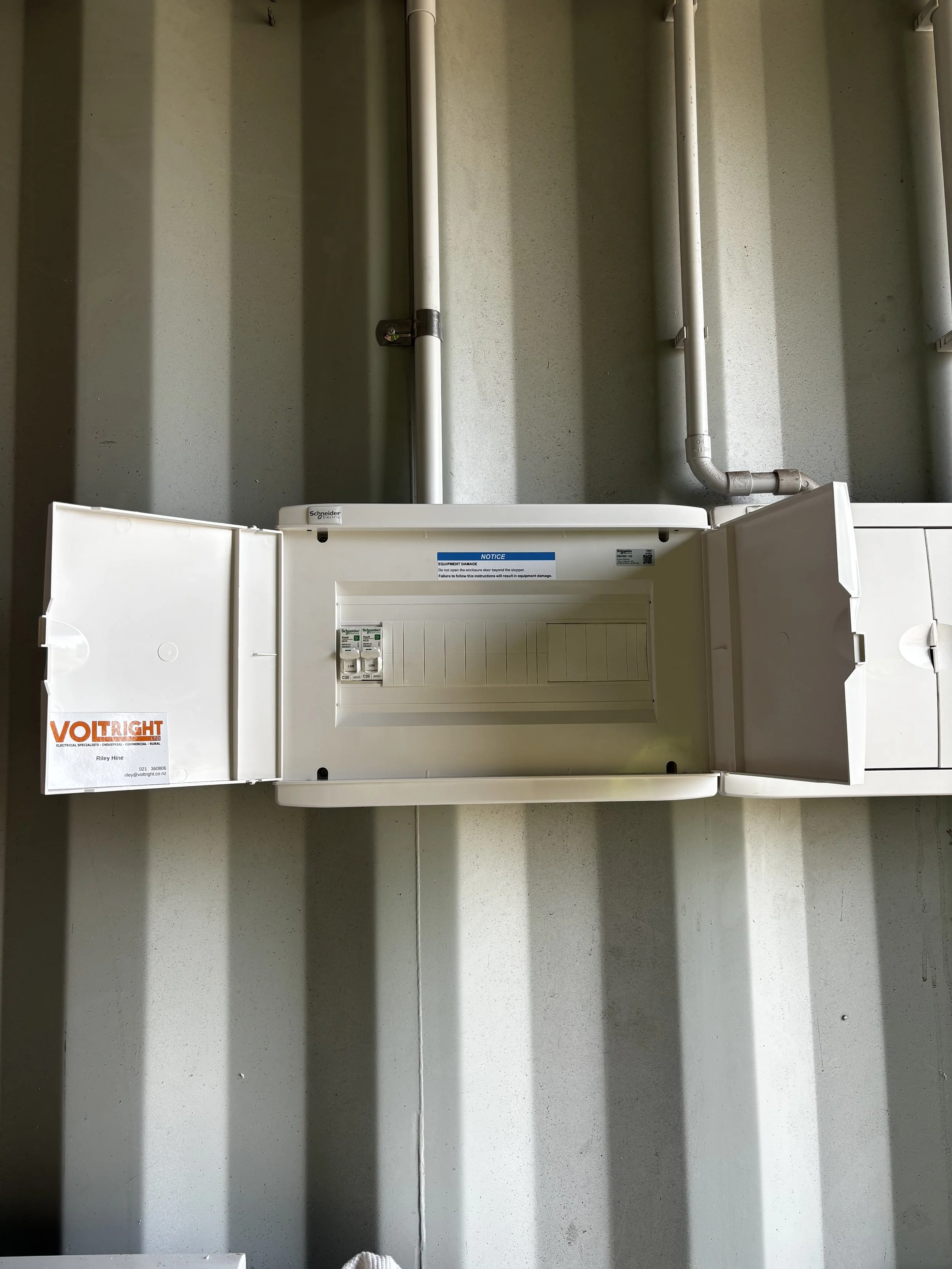 Interior of an electrical breaker box with open door, mounted on a wall with gray vertical lines, and white pipes running vertically behind it.