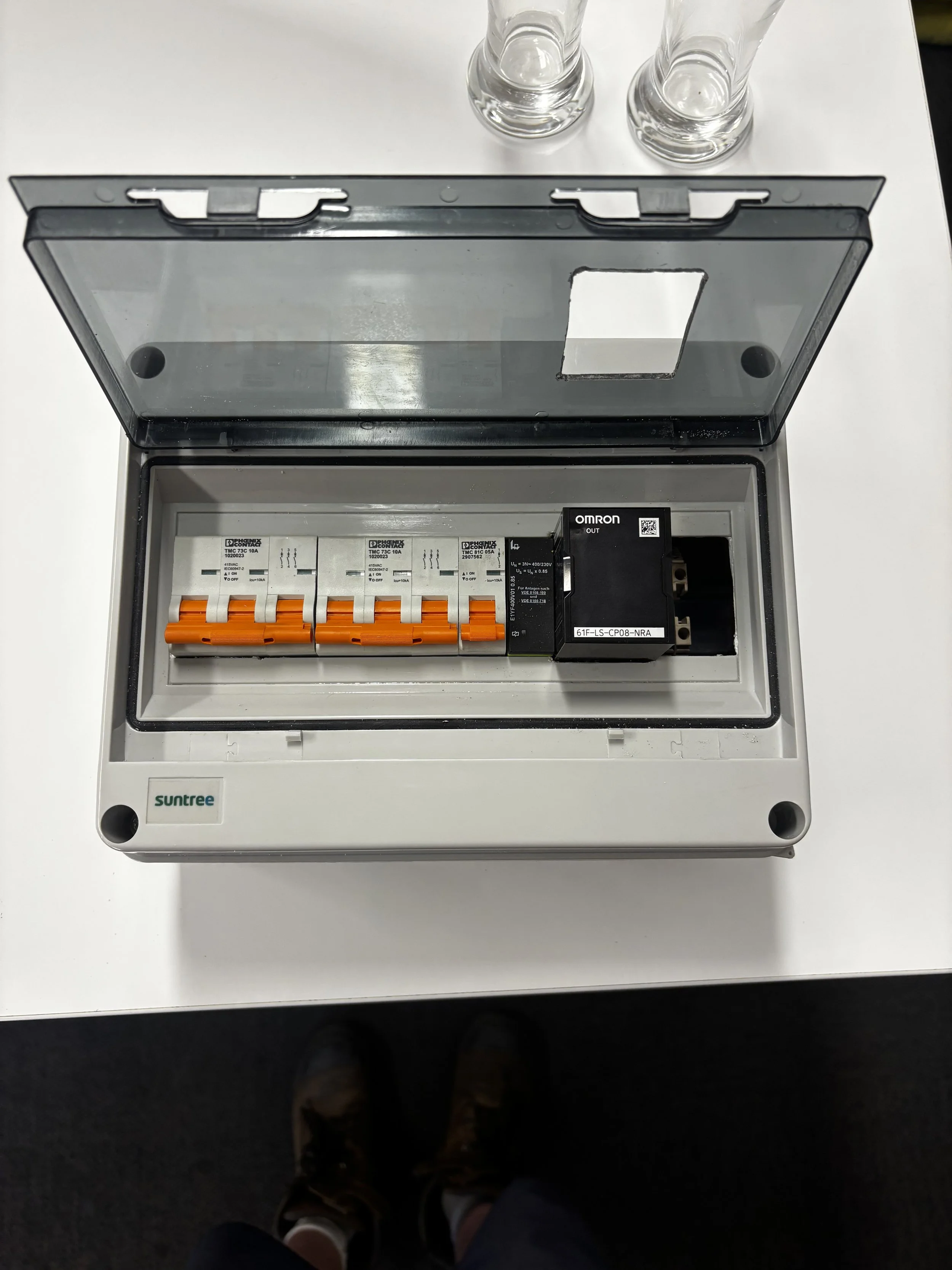 An electrical circuit breaker box with four switches and an Omron relay inside, with a transparent cover, on a white table with two empty glass bottles.