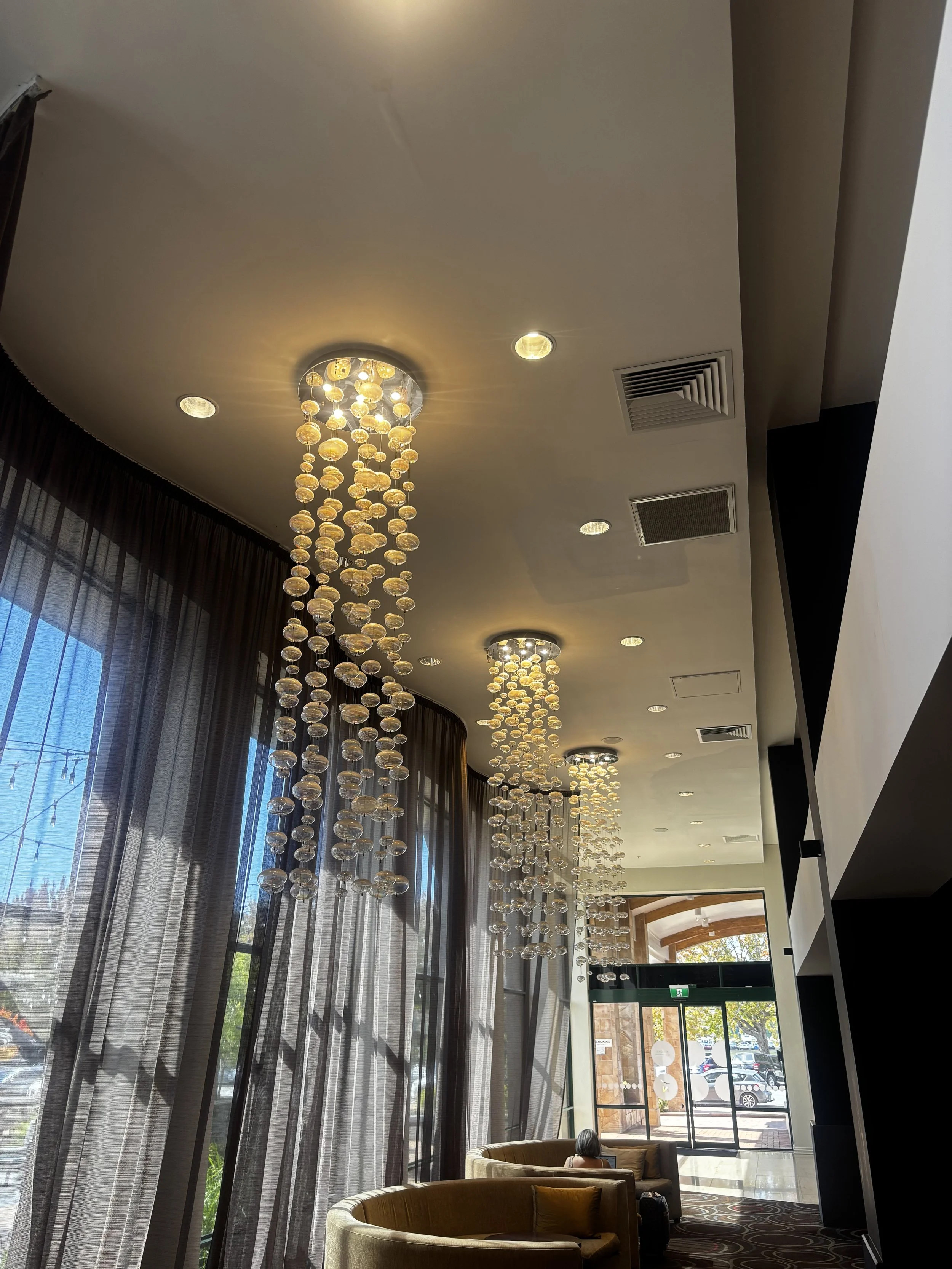Interior of a modern lobby or lounge area with tall windows, sheer curtains, and contemporary chandeliers with hanging glass orbs, with a person seated on a circular sofa near the entrance.