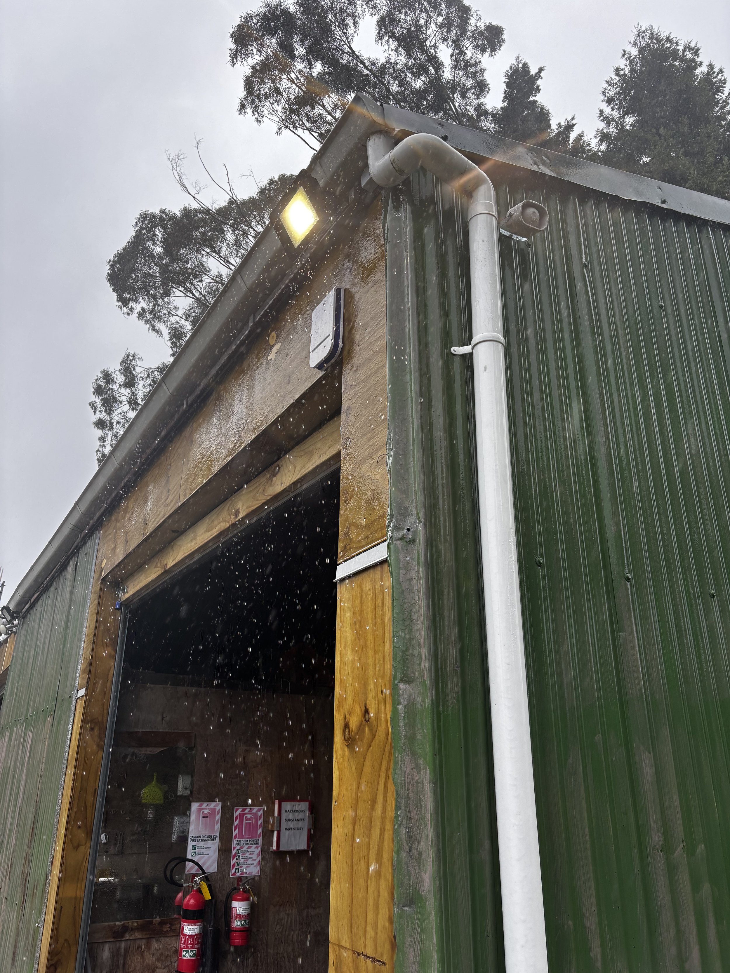 Corner of a green metal building with a white vertical pipe, an outdoor light, and security camera, with trees in the background during rainy weather.