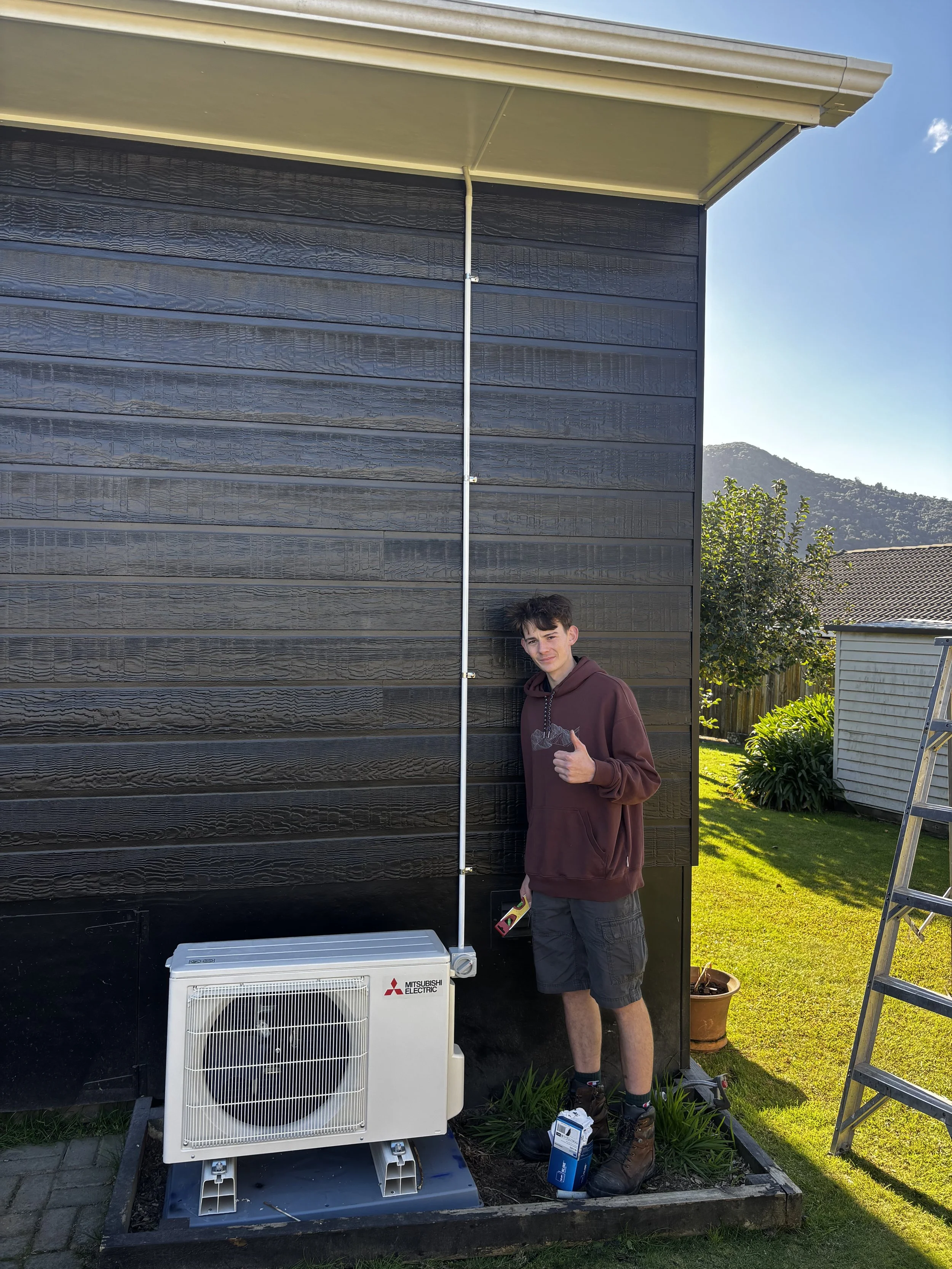 A young man in a brown hoodie and black shorts giving a thumbs-up sign while standing next to an outdoor air conditioning unit on a landscaped patch beside a black house with horizontal siding. There is a ladder and a potted plant nearby, and a mount