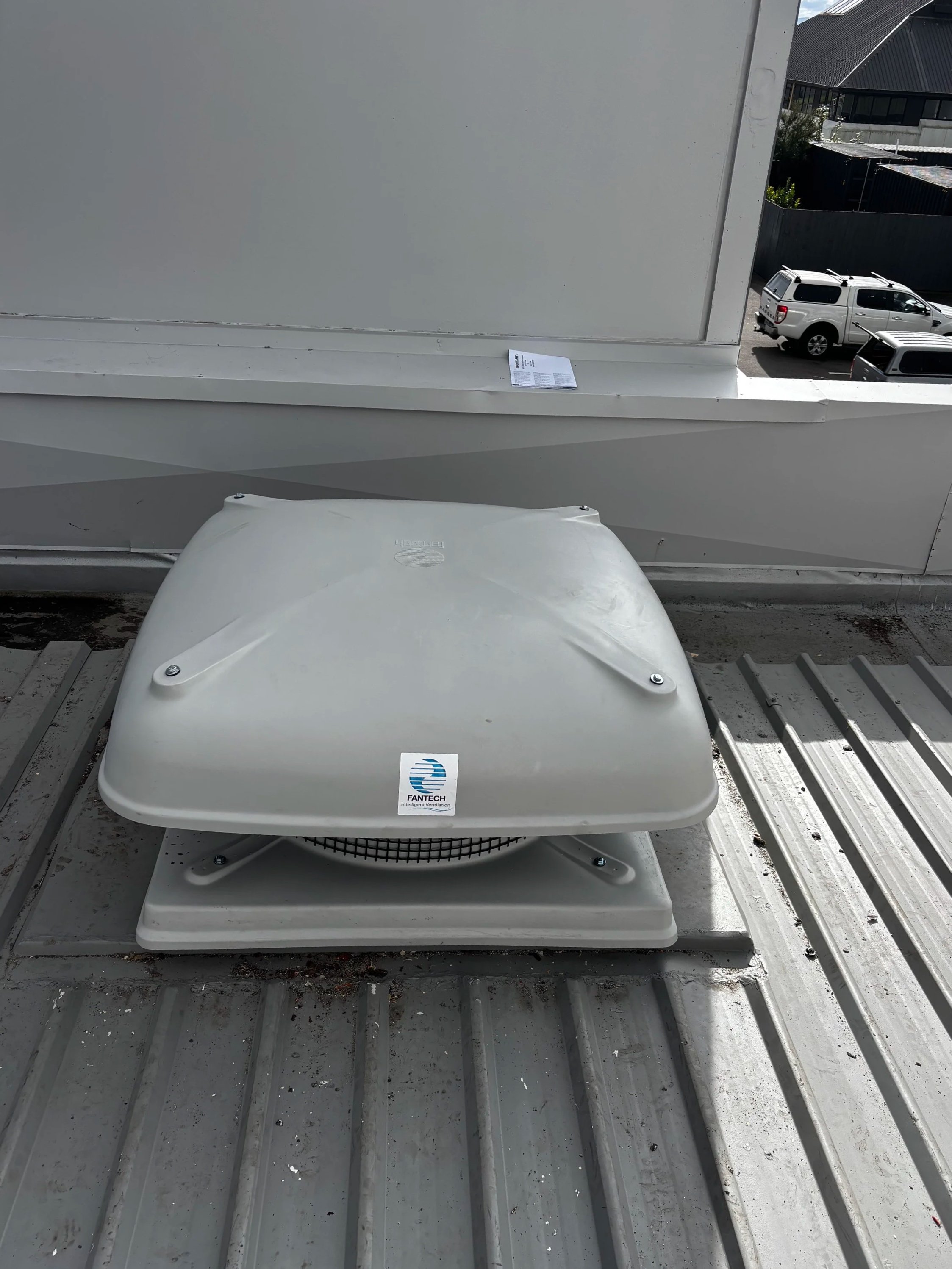 An industrial ventilation fan installed on a metal rooftop, with a shipping label on the top of the fan and another on the wall behind it.