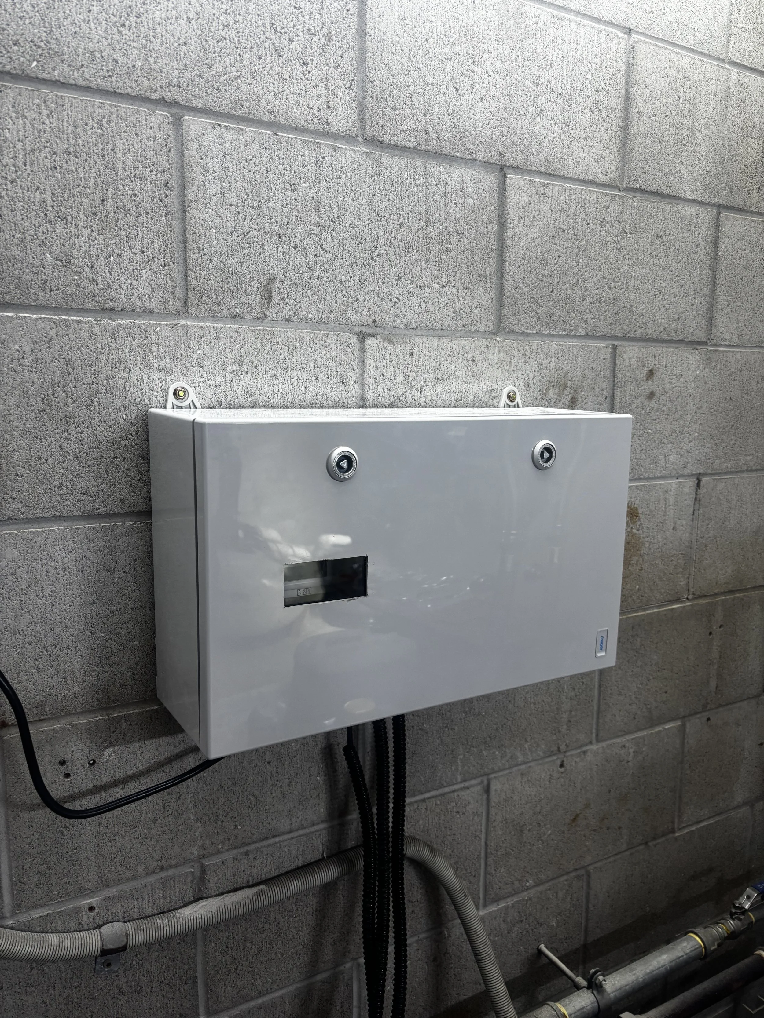 A gray electrical panel mounted on a concrete wall with visible wiring and pipes below it.