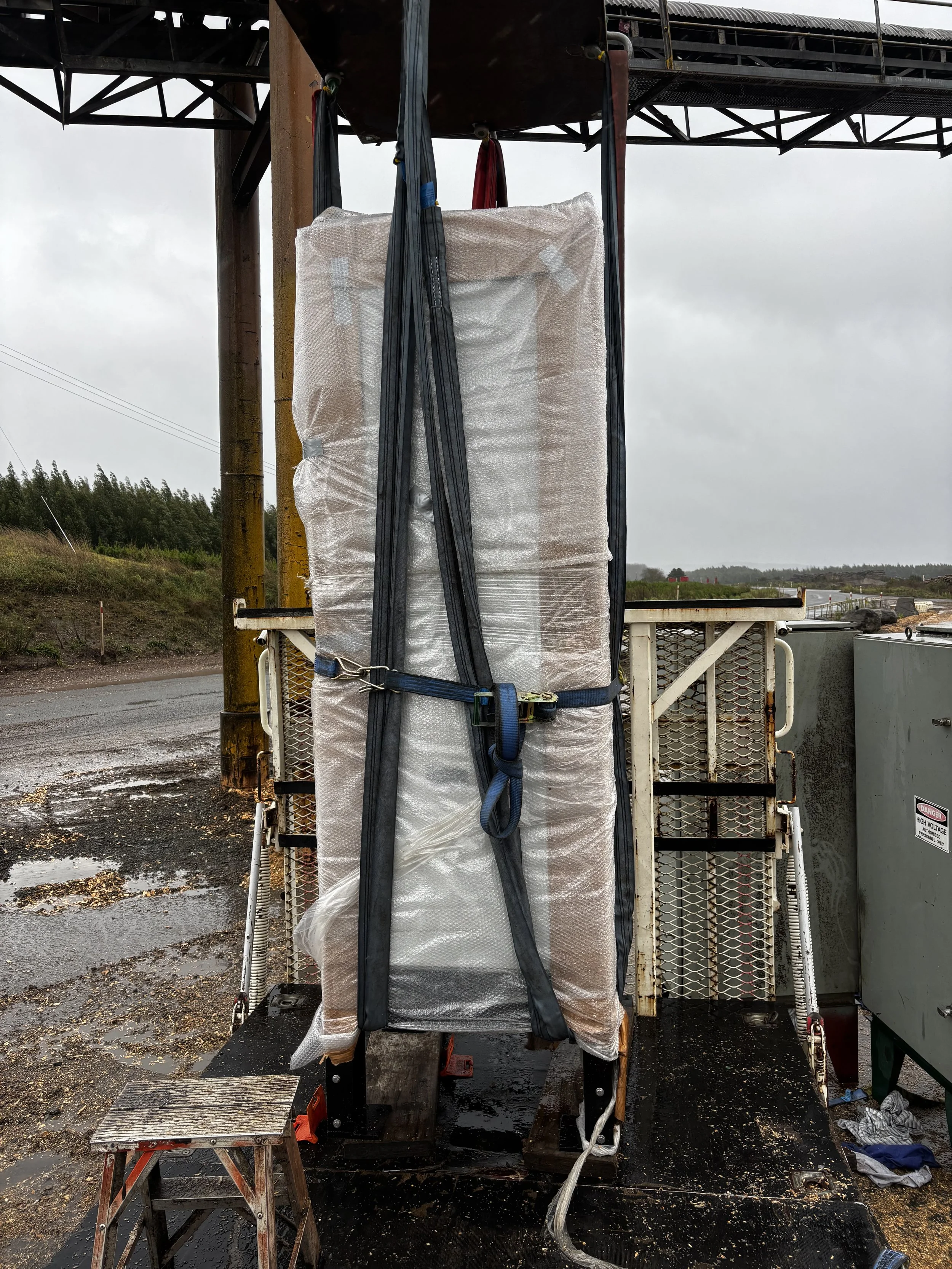Large wrapped pallet secured with black straps on industrial equipment, outdoors on wet ground with cloudy sky.