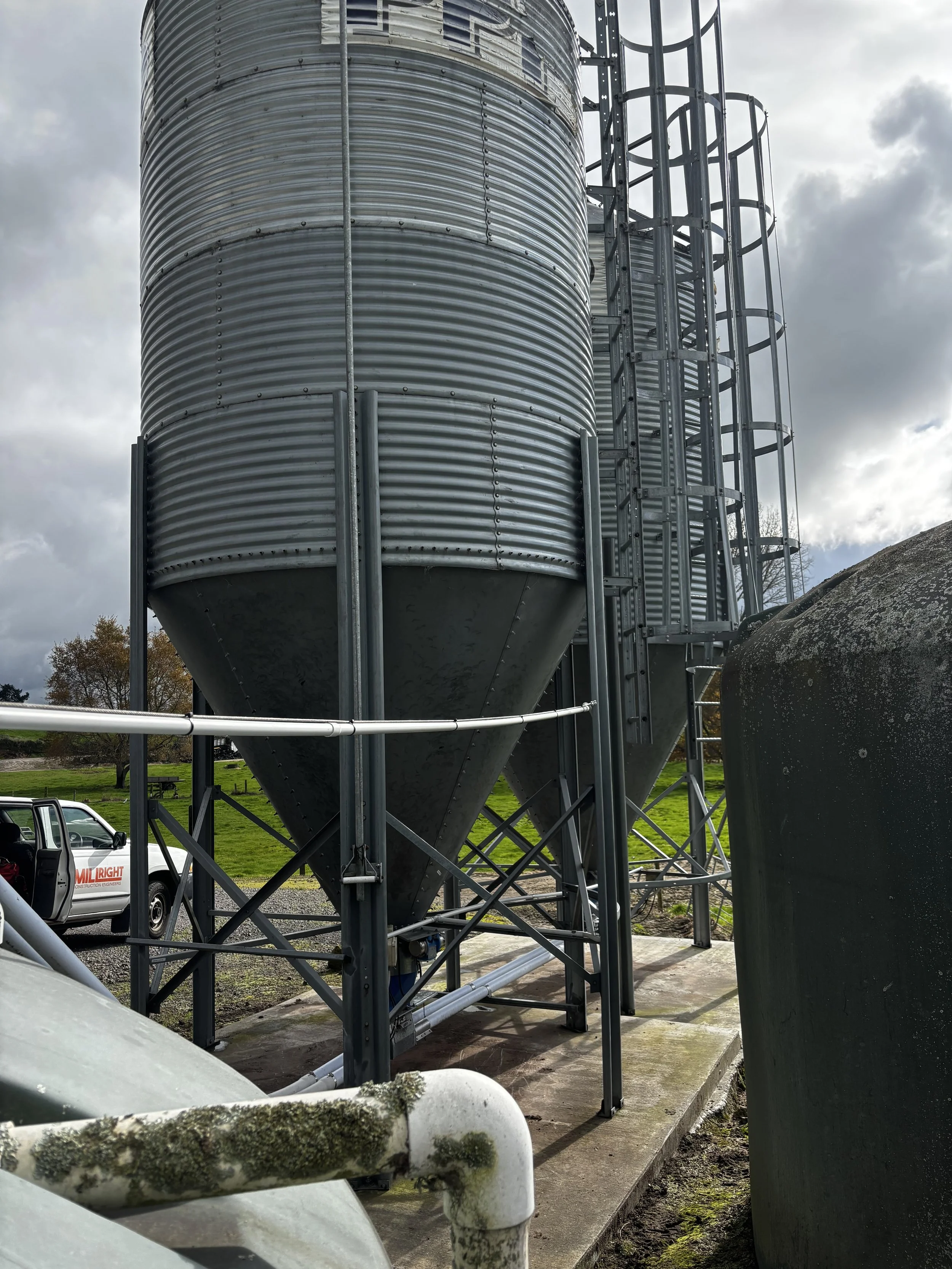 A large, metallic, cylindrical storage silo with a conical bottom, situated outdoors on a concrete pad, with metal stairs and ladders attached, under a partly cloudy sky, with some greenery and an industrial van nearby.