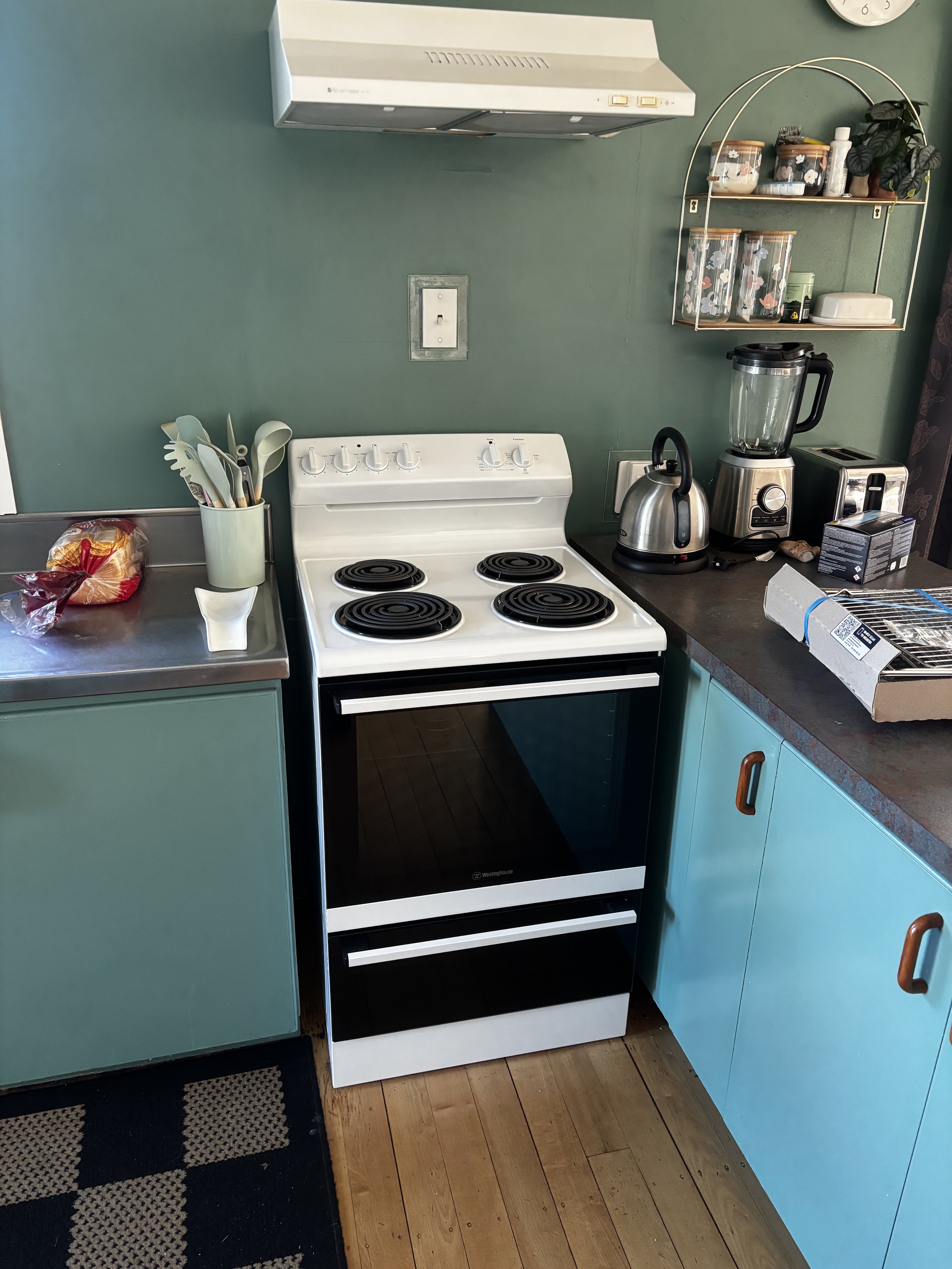 Kitchen with a vintage white electric stove, black oven, turquoise cabinets, a small countertop with a bag of bread, utensil holder, toaster, blender, kettle, and spice jars, with a wall shelf and a clock.